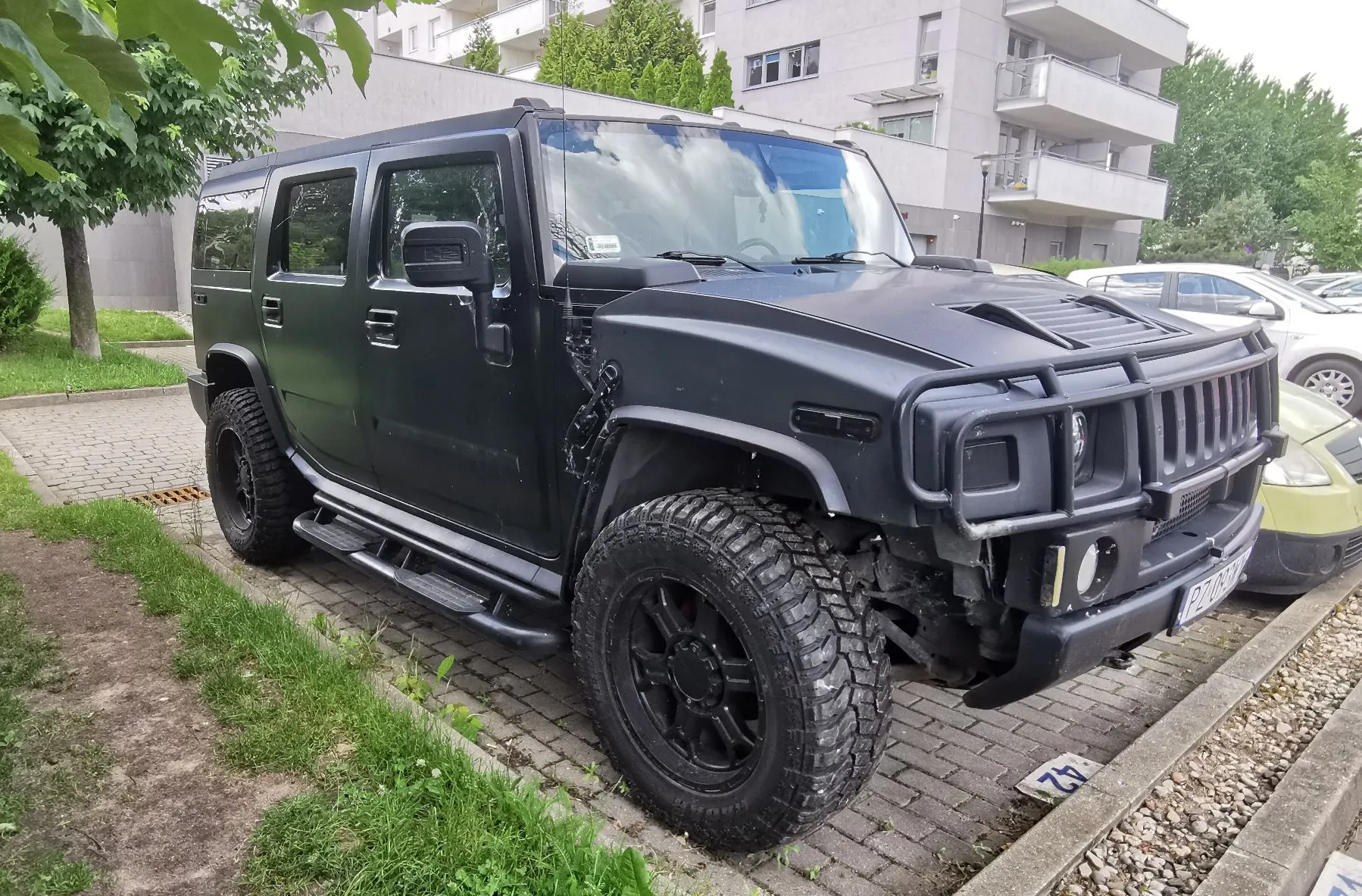 Czarny, matowy samochód terenowy typu Hummer zaparkowany na kostce brukowej obok trawnika. Pojazd posiada duże, terenowe opony, czarne felgi oraz masywny zderzak z osłoną grillu. W tle widoczny jest nowoczesny, jasny budynek mieszkalny z balkonami oraz inne zaparkowane samochody.