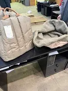 Wnętrze sklepu z czarnym stołem, na którym leży beżowa pikowana kurtka z białą etykietą oraz szara tkanina. Obok stołu znajduje się czarna szafka z szufladami. W tle widoczne są półki z różnymi przedmiotami i betonowa podłoga.