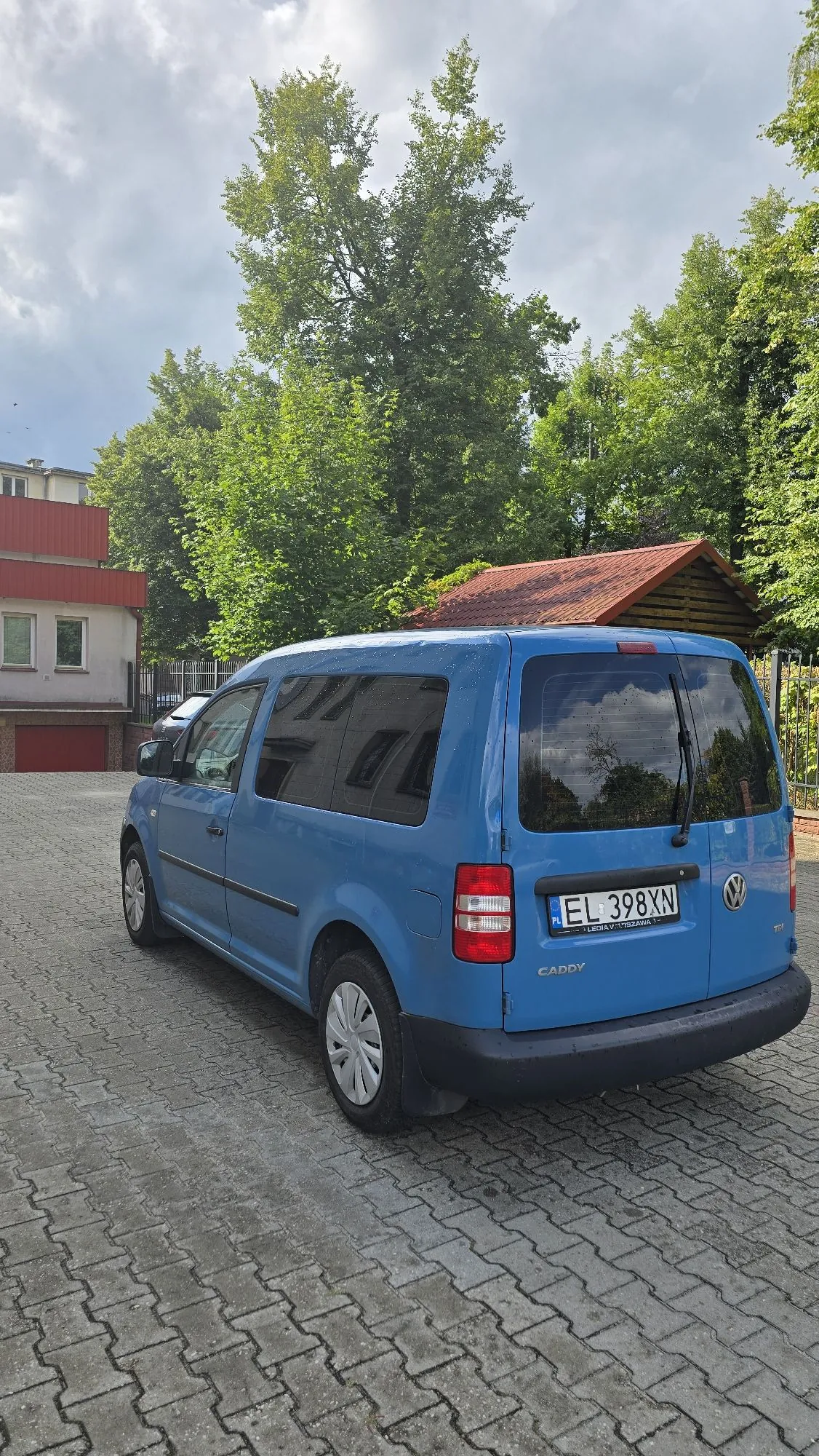 Niebieski samochód dostawczy Volkswagen Caddy zaparkowany tyłem na kostce brukowej. Pojazd ma polską tablicę rejestracyjną o numerze EL 398XN. W tle widoczne są zielone drzewa, fragment czerwonego budynku oraz drewniany dach altany pod zachmurzonym niebem.