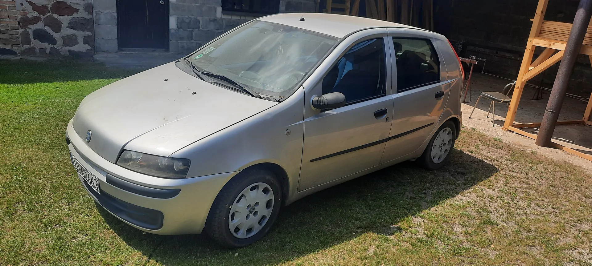 Srebrny samochód osobowy typu hatchback zaparkowany na trawie. Widoczna tablica rejestracyjna z numerem WU 6763. W tle kamienica z cegły i betonu z czarnymi drzwiami, drewniana konstrukcja oraz krzesło. Trawa jest zielona, a na ziemi widoczne są cienie od słońca.