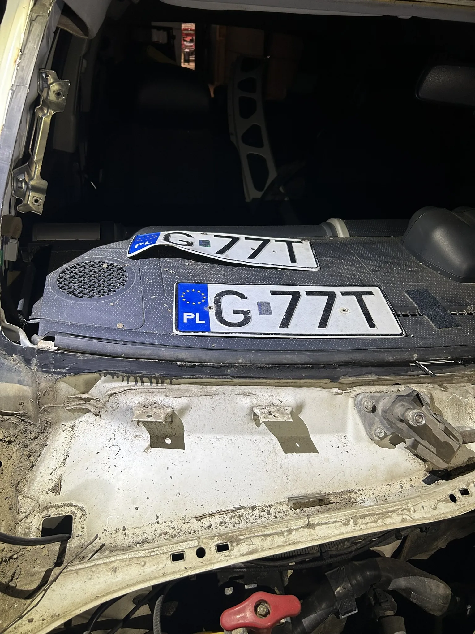 Wnętrze uszkodzonego samochodu z widoczną deską rozdzielczą. Na desce znajdują się dwie tablice rejestracyjne z napisem 'G-77T' oraz niebieskim polem z napisem 'PL' i symbolem Unii Europejskiej. Widoczne są również uszkodzone elementy karoserii, czarna siatka głośnika oraz części silnika na dole kadru. Tło jest ciemne, sugerujące wnętrze garażu lub ciemnego pomieszczenia.