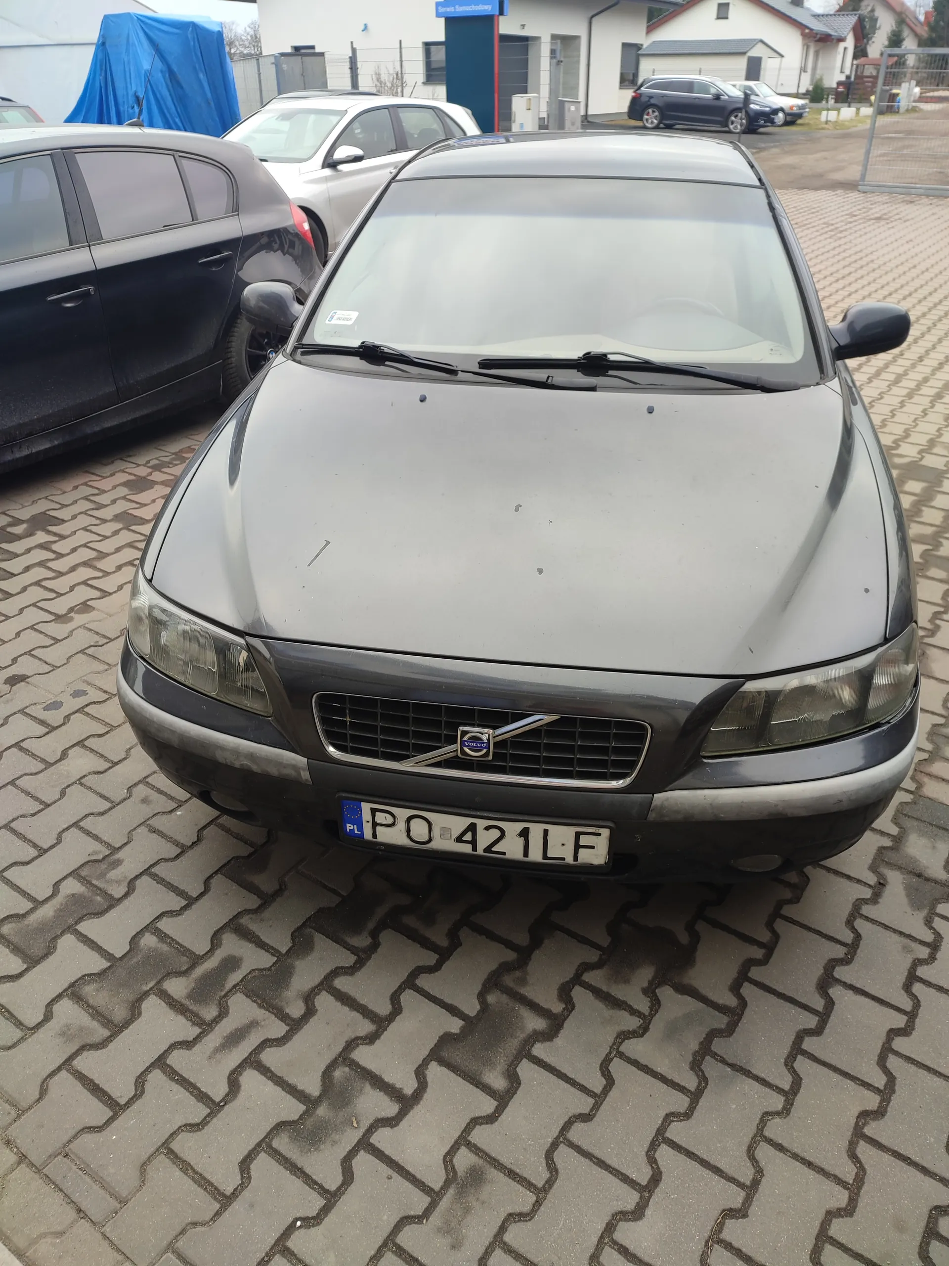 Przedni widok szarego samochodu marki Volvo z tablicą rejestracyjną PO 421 LF (PL) zaparkowanego na brukowanej powierzchni. W tle widoczne są inne pojazdy, budynek z niebieskim napisem oraz niebieska folia przykrywająca przedmiot. Samochód ma czystą szybę przednią i charakterystyczny emblemat Volvo na masce.