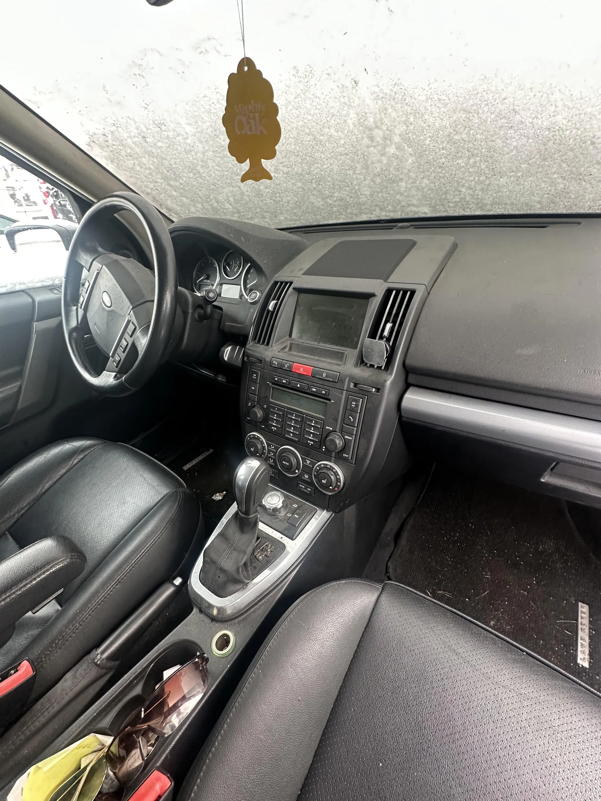 Wnętrze samochodu z widoczną czarną kierownicą po lewej stronie, tablicą rozdzielczą z licznikami, centralnym panelem z radioodtwarzaczem, przyciskami i gałkami sterującymi. Na desce rozdzielczej wiszący aromatyzator w kształcie drzewa z napisem "Mighty Oak". Czarne skórzane fotele, dźwignia zmiany biegów oraz przedmioty w uchwycie na kubki (m.in. okulary przeciwsłoneczne i żółty obiekt). Przednią szybę pokrywa szron. Podłoga wewnątrz pojazdu jest zabrudzona.