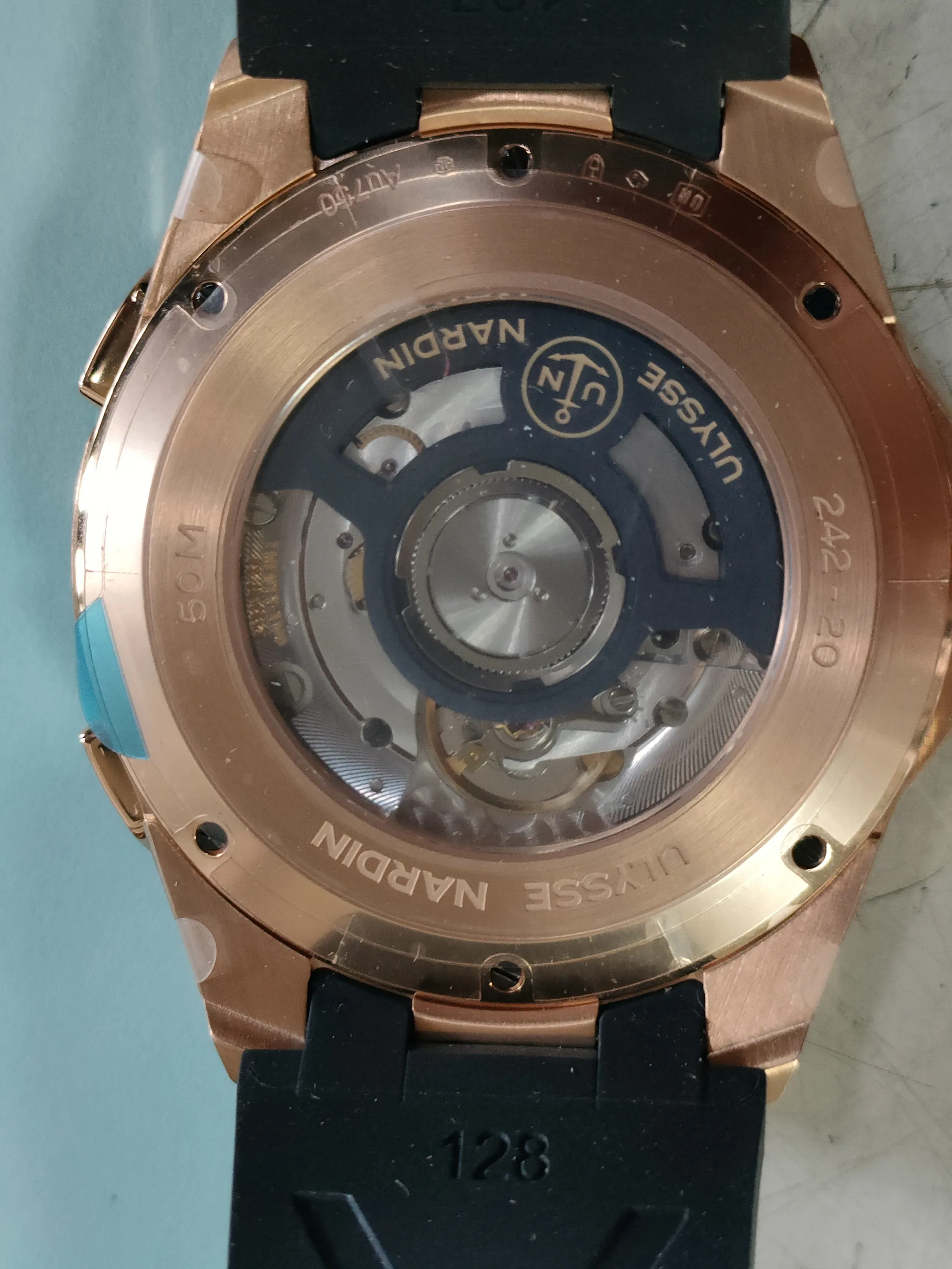 Zbliżenie na tylną część zegarka z przezroczystą kopertą pokazującą mechanizm. Obudowa w kolorze różowego złota z napisem 'ULYSSE NARDIN' wokół krawędzi, logo z kotwicą i literami 'UN', oznaczenie '50M' oraz cyfry '242-20'. Czarna gumowa bransoleta z widocznym numerem '128'. Tło w jasnoniebieskim kolorze.