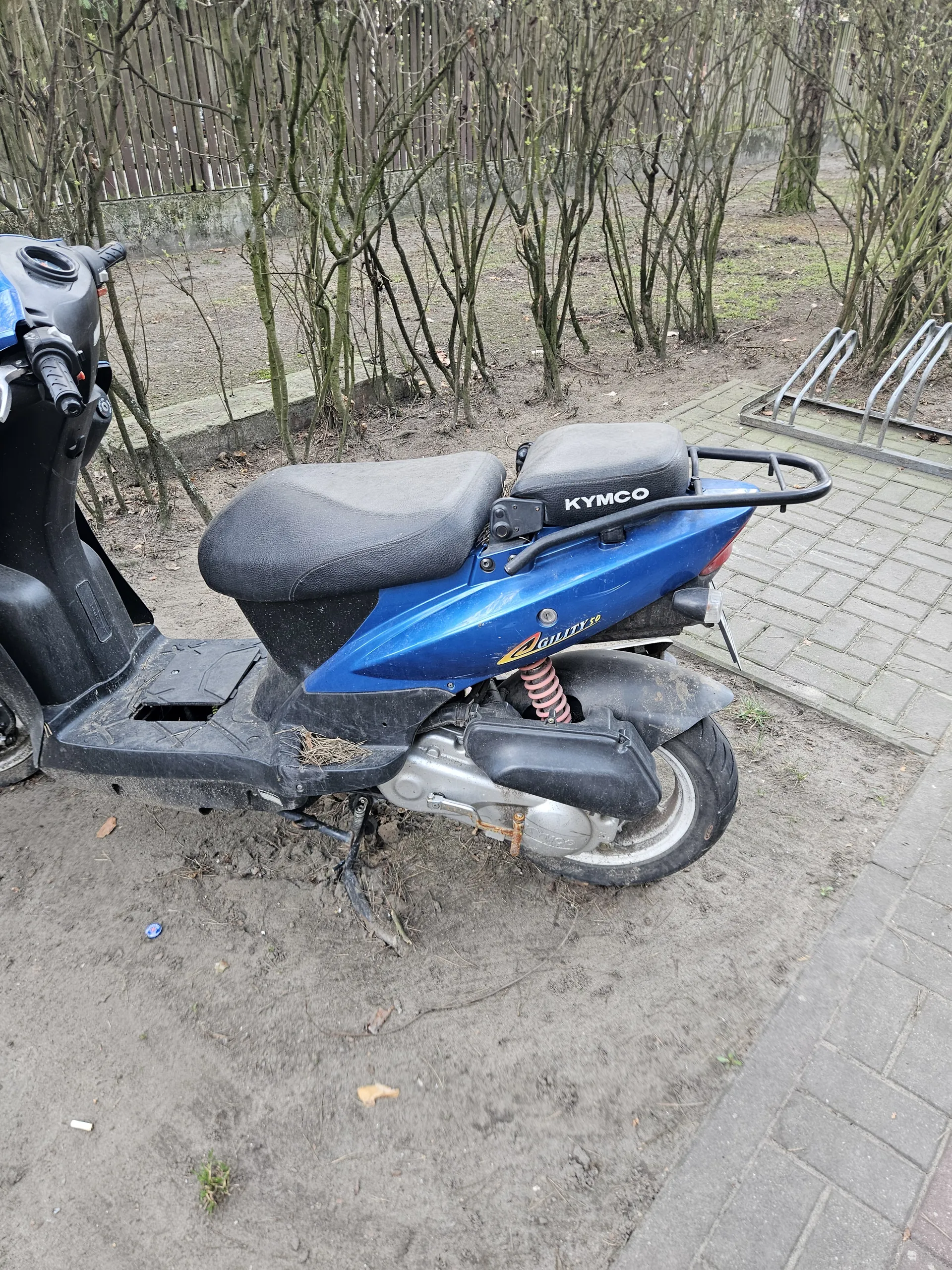 Niebieski skuter marki Kymco z napisem 'KYMCO' na siedzeniu i 'AGILITY' na boku, zaparkowany na brudnej ziemi obok chodnika z cegieł. W tle znajdują się nagie krzewy i metalowy stojak na rowery. Na ziemi widoczne są rozrzucone liście i małe przedmioty.