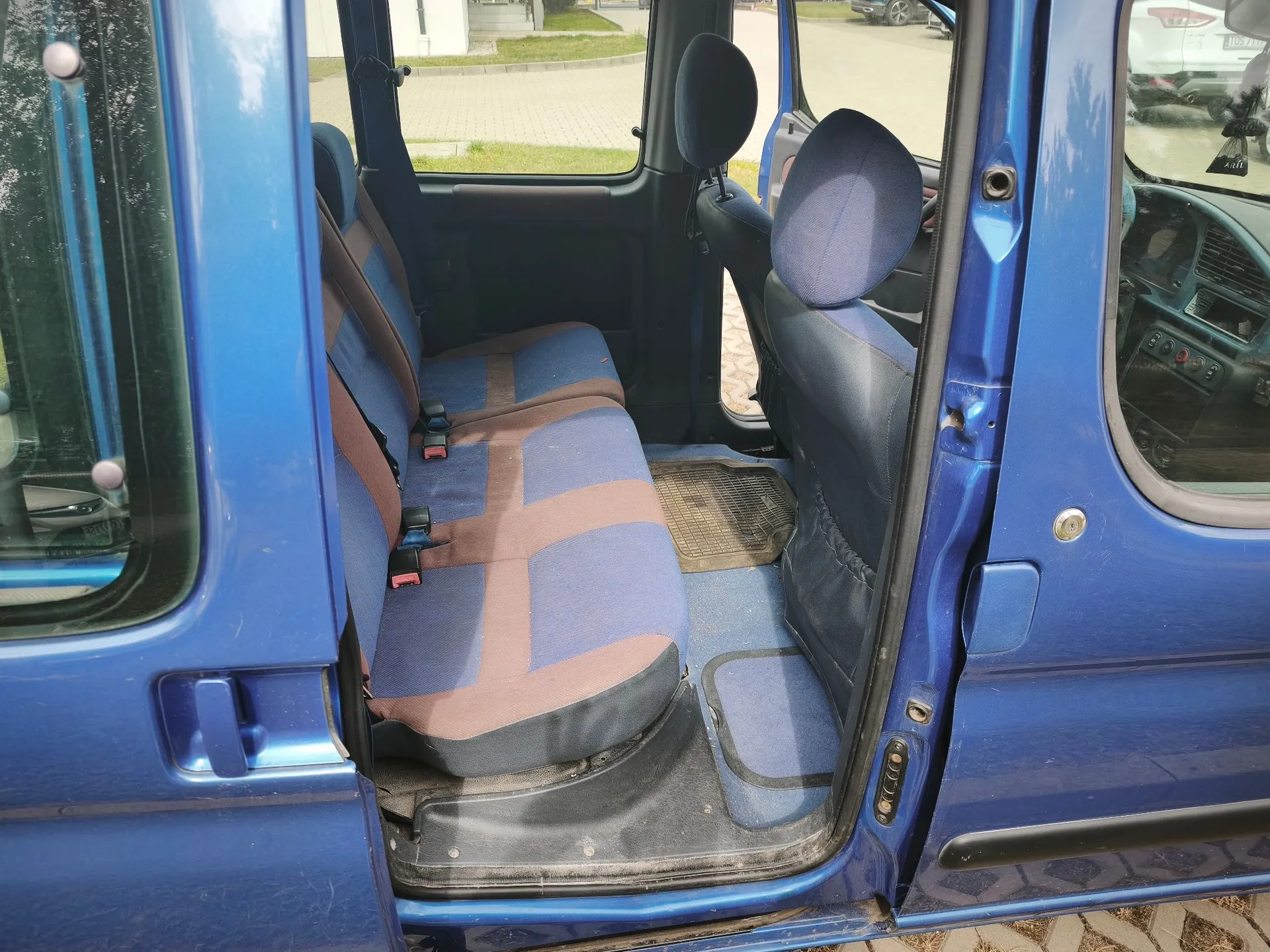 Widok z wnętrza niebieskiego samochodu przez otwarte drzwi pasażera. Widoczna jest tylna kanapa z niebiesko-brązową tapicerką w kwadraty oraz przedni fotel z zagłówkiem. Podłoga jest wyłożona niebieskimi gumowymi dywanikami.