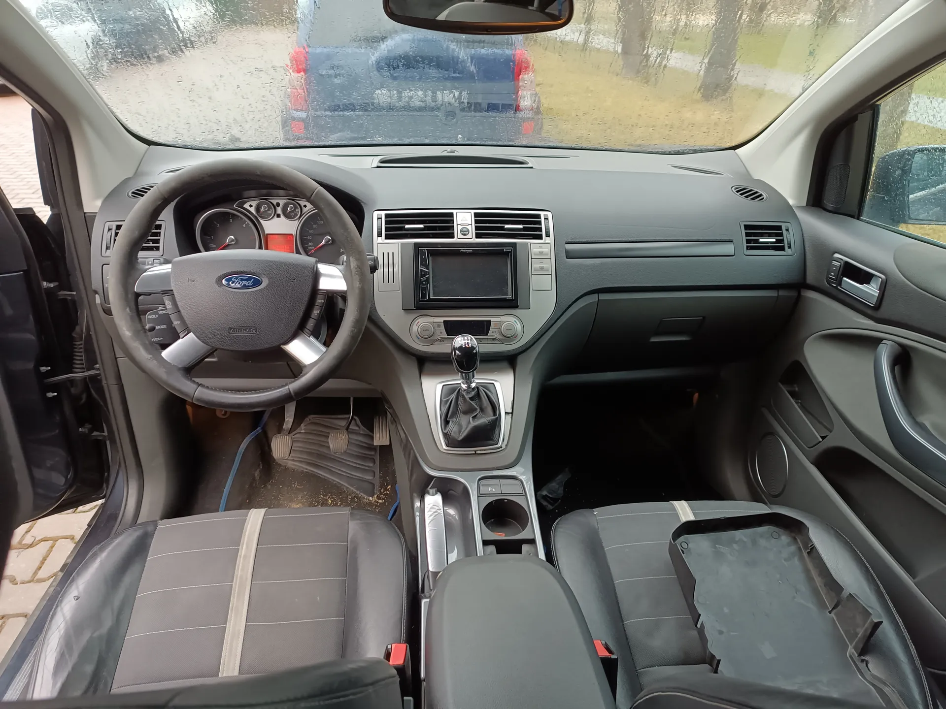 Wnętrze samochodu marki Ford z widocznym kierownicą z logo Ford, deska rozdzielcza z ekranem, przyciskami i wentylacjami, dźwignią zmiany biegów oraz siedzeniami w kolorze szarym z pionowymi paskami. Drzwi po stronie kierowcy są otwarte, przez przednią szybę widać niebieski samochód Suzuki z czytelnym napisem "SUZUKI" na tylnej szybie. Na przedniej szybie znajdują się krople deszczu, a poza samochodem widoczne są drzewa i trawnik. Wnętrze posiada czarne i szare elementy, a na podłodze widoczne są ślady brudu.