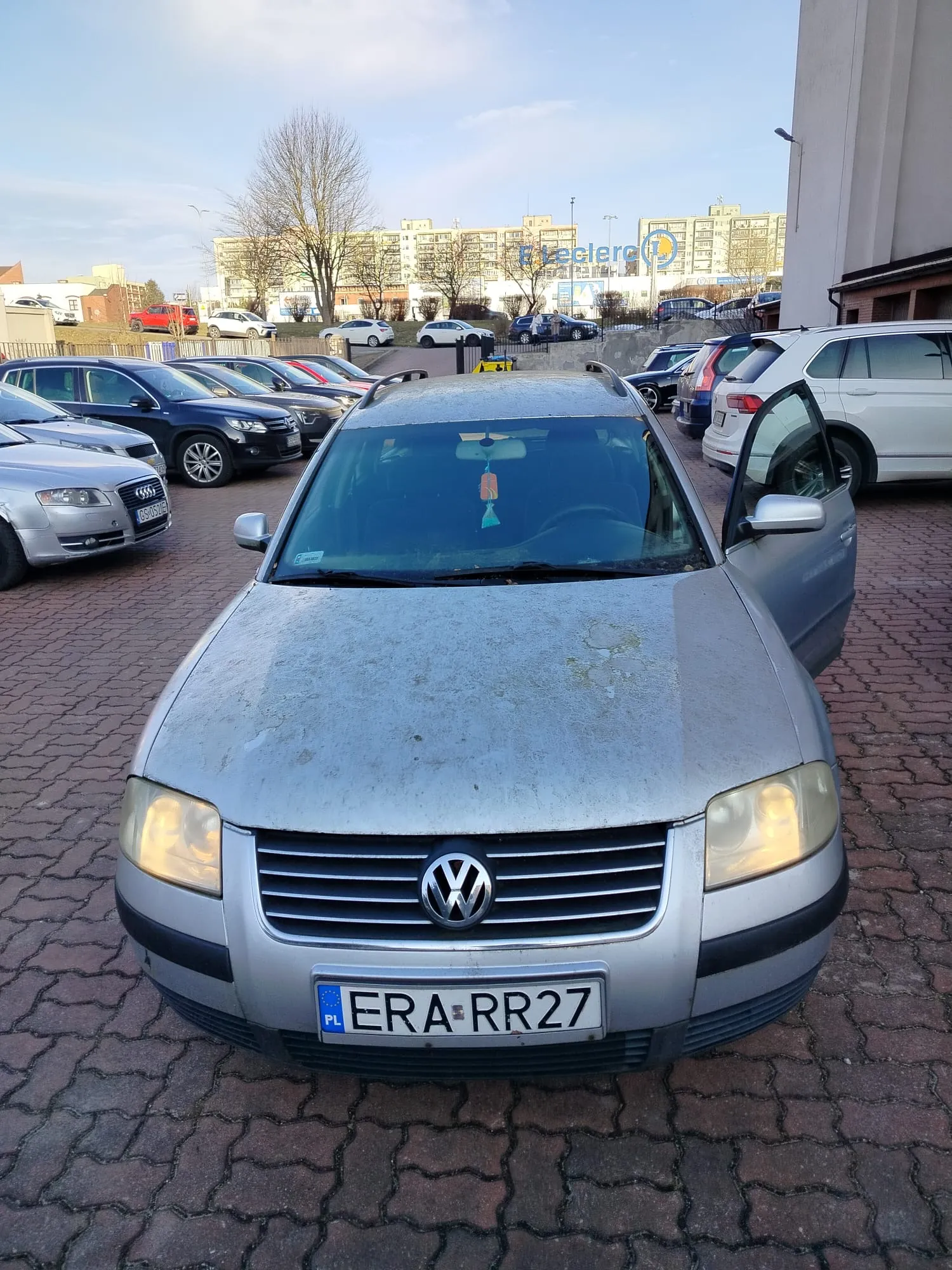 Srebrny samochód marki Volkswagen z tablicą rejestracyjną ERA RR27 (PL) zaparkowany na brukowanym parkingu. Drzwi pasażera po lewej stronie są otwarte. W tle widoczne są inne zaparkowane pojazdy, budynki mieszkalne oraz sklep E.Leclerc. Na przedniej szybie wiszą ozdoby. Niebo jest częściowo zachmurzone.