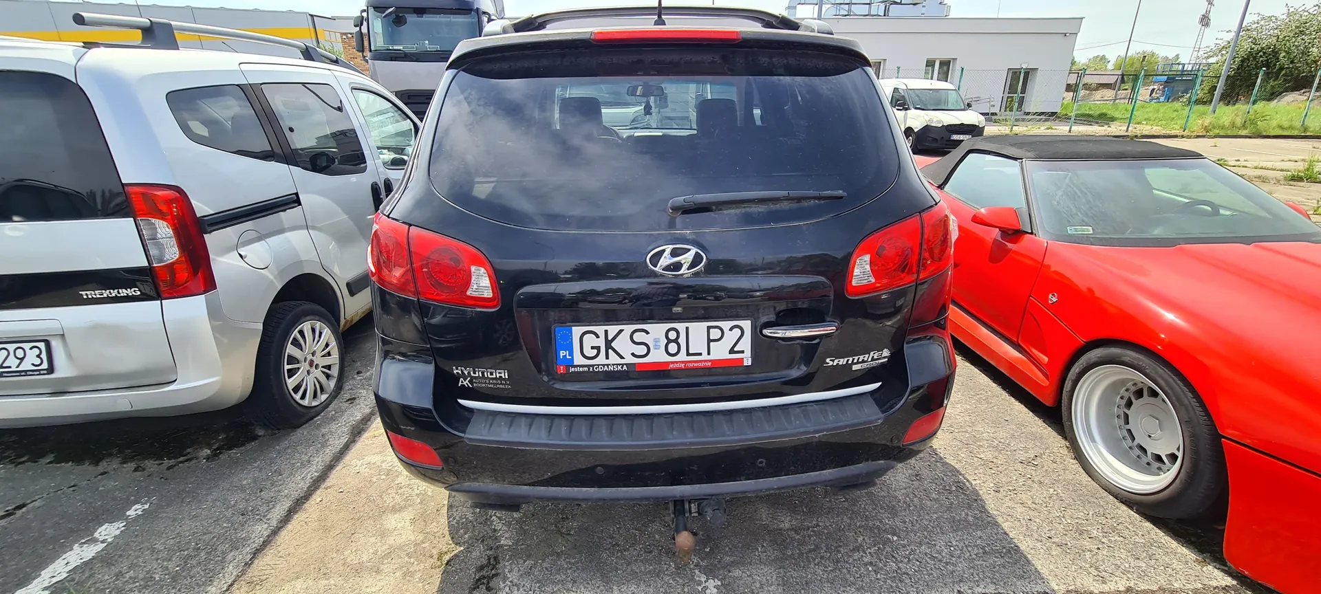 Zdjęcie przedstawia czarny samochód Hyundai Santa Fe z tablicą rejestracyjną PL GKS 8LP2, zaparkowany na placu parkingowym. Po lewej stronie widoczny srebrny van z napisem TREKKING i częściowo widoczną tablicą 293. Po prawej stronie czerwony kabriolet. Na tle budynek, ogrodzenie, zielona trawa oraz inne pojazdy. Na tablicy rejestracyjnej czarnego Hyundaia widoczny napis 'jestem z GDAŃSKA' na czerwonym tle.