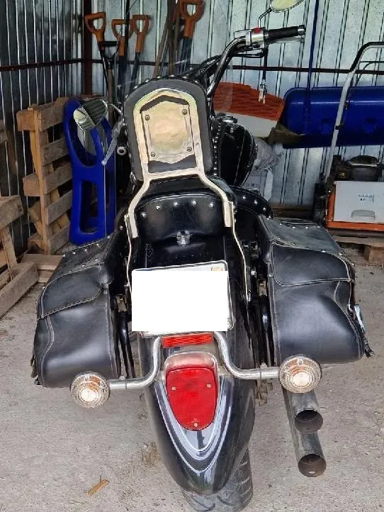 Widok z tyłu na czarny motocykl typu cruiser z dwiema skórzanymi sakwami po bokach, oparciem dla pasażera oraz chromowanymi rurami wydechowymi i lampami. Motocykl znajduje się w garażu lub szopie na tle metalowej ściany i drewnianych palet.