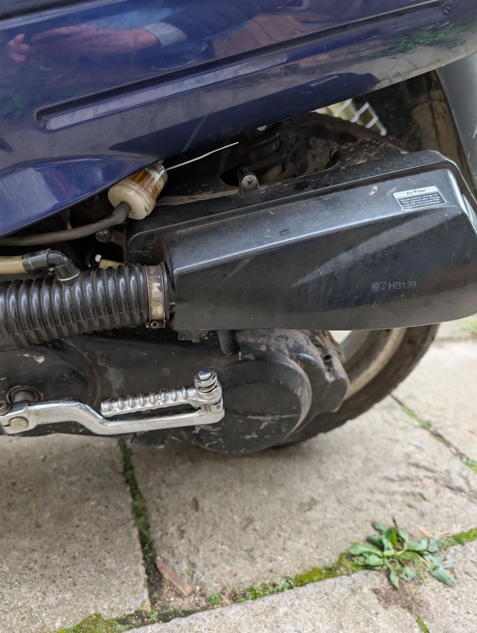 Zbliżenie na dolną część niebieskiego skutera. Widoczna jest czarna obudowa filtra powietrza z naklejką zawierającą tekst 'Air Filter' oraz oznaczeniem 'HB139'. Poniżej znajduje się metalowa stopka boczna oraz elementy układu napędowego. W tle widoczne jest koło skutera i betonowe płyty chodnikowe.