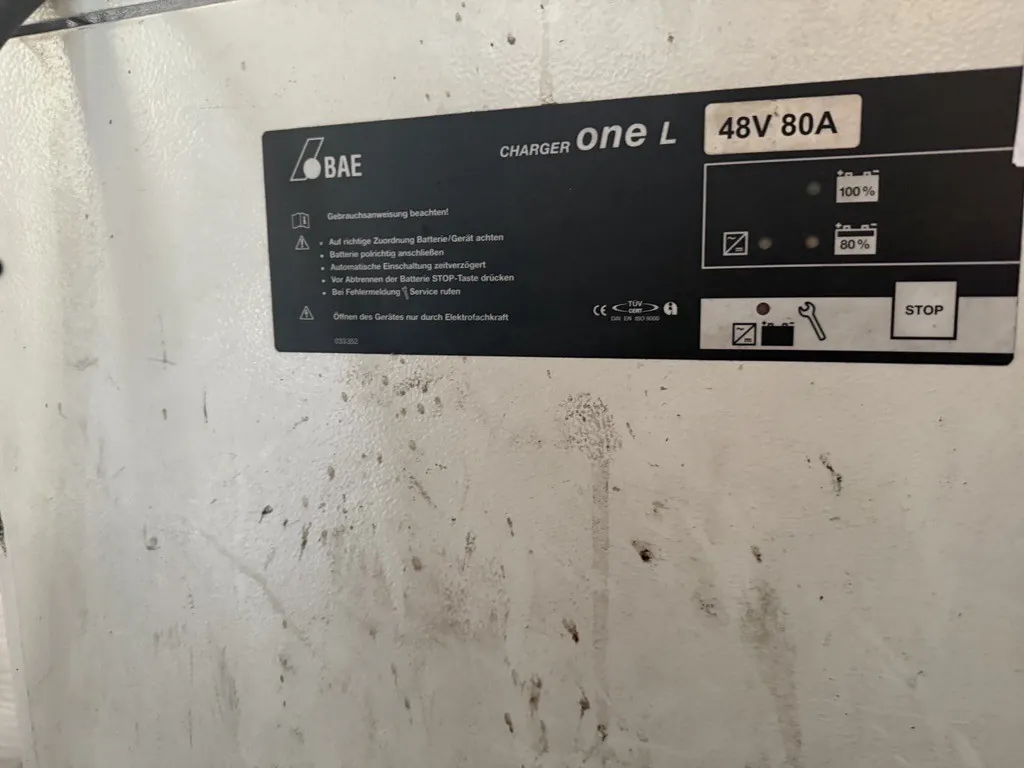 Zdjęcie tabliczki znamionowej ładowarki marki BAE. Na czarnym tle widnieje biały napis 'CHARGER One L 48V 80A'. Poniżej znajduje się instrukcja w języku niemieckim z ostrzeżeniami, m.in. 'Gebrauchsanweisung beachten!'. Po prawej stronie znajdują się ikony poziomu naładowania baterii (100% i 80%), symbol klucza serwisowego oraz przycisk z napisem 'STOP'. W dolnej części widoczne są certyfikaty CE i TÜV.