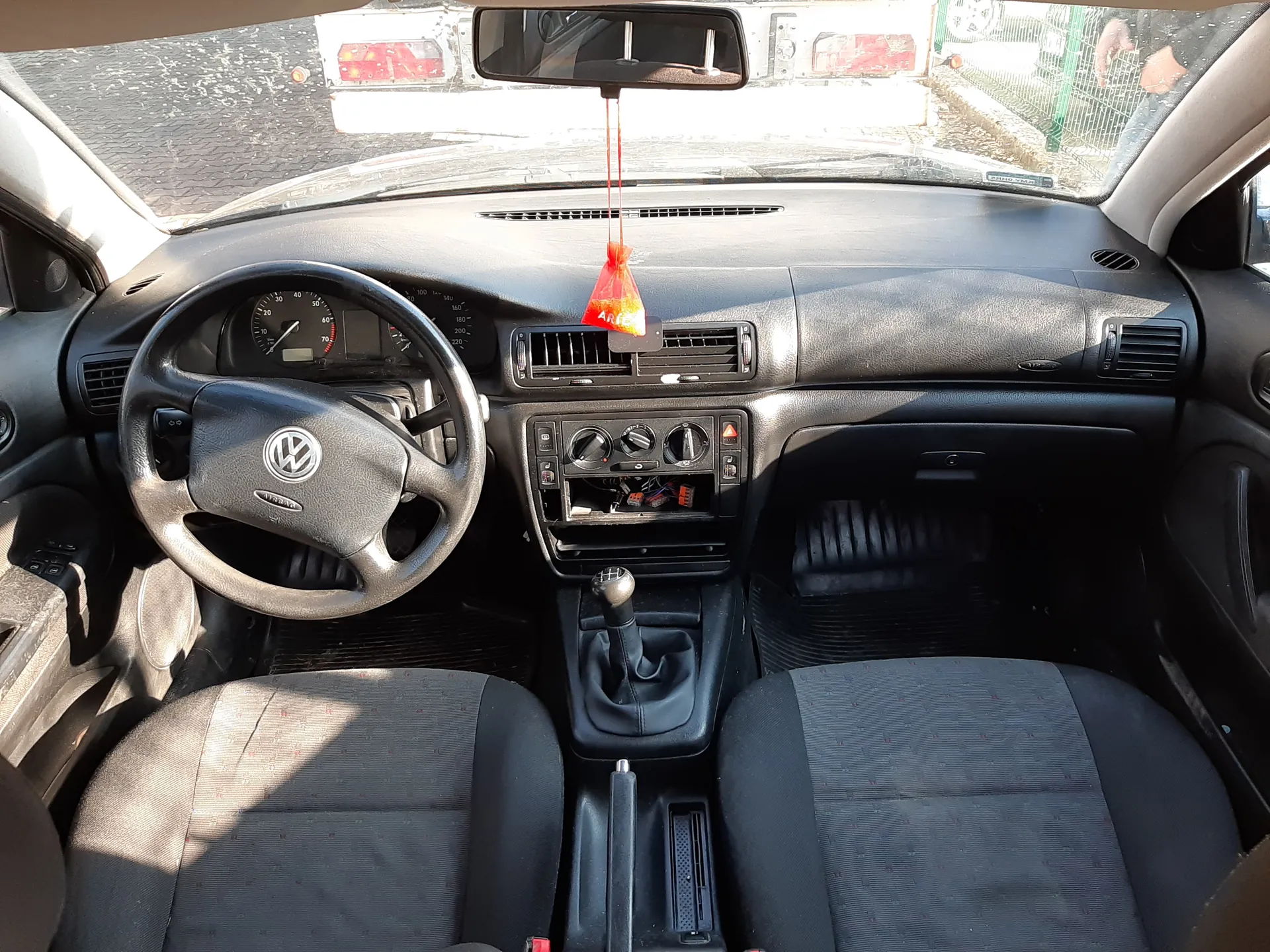 Wnętrze samochodu Volkswagen z czarną deską rozdzielczą, kierownicą z logo VW, widocznym licznikiem prędkości, dźwignią zmiany biegów i przednimi siedzeniami w szaro-czarnym kolorze. Na lusterku wstecznym wisząca czerwona ozdoba. W centralnej konsoli otwarta komora z odsłoniętymi przewodami. Przez przednią szybę widoczny inny pojazd i ogrodzenie.