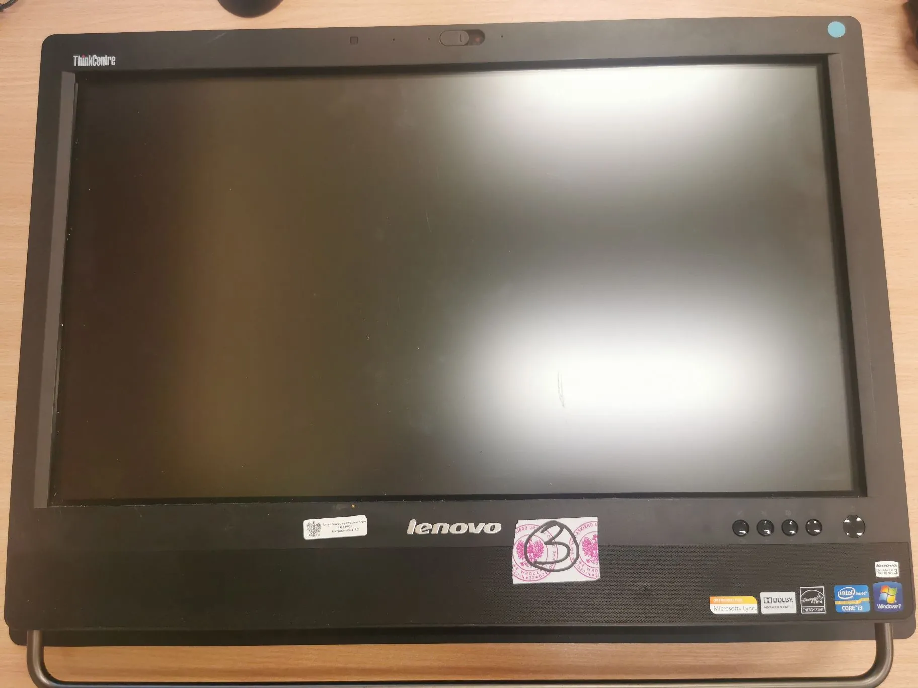 Czarny monitor marki Lenovo ThinkCentre z wyłączonym ekranem. Na ramce na górze widnieje napis 'ThinkCentre', a na dole centralnie logo 'lenovo'. Poniżej logo znajdują się naklejki informacyjne, w tym logo Intel Core i3 oraz Windows 7. Po prawej stronie dolnej ramki znajdują się przyciski sterujące.