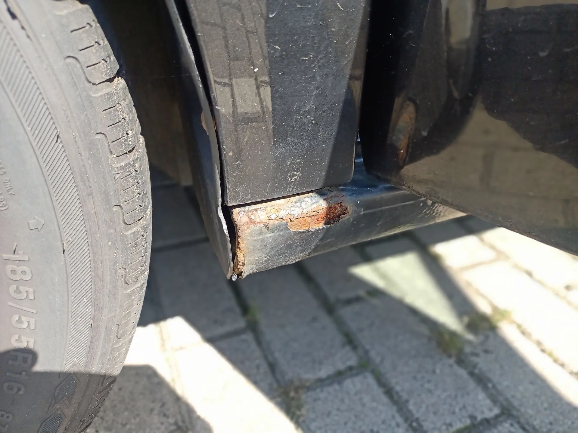 Zbliżenie na dolną część czarnego nadwozia samochodu w pobliżu przedniego koła. Metalowy element progu jest silnie skorodowany, z widocznymi ubytkami w materiale i rdzawym nalotem. Po lewej stronie widoczny jest fragment opony z oznaczeniem rozmiaru 185/55 R16. Samochód stoi na kostce brukowej.