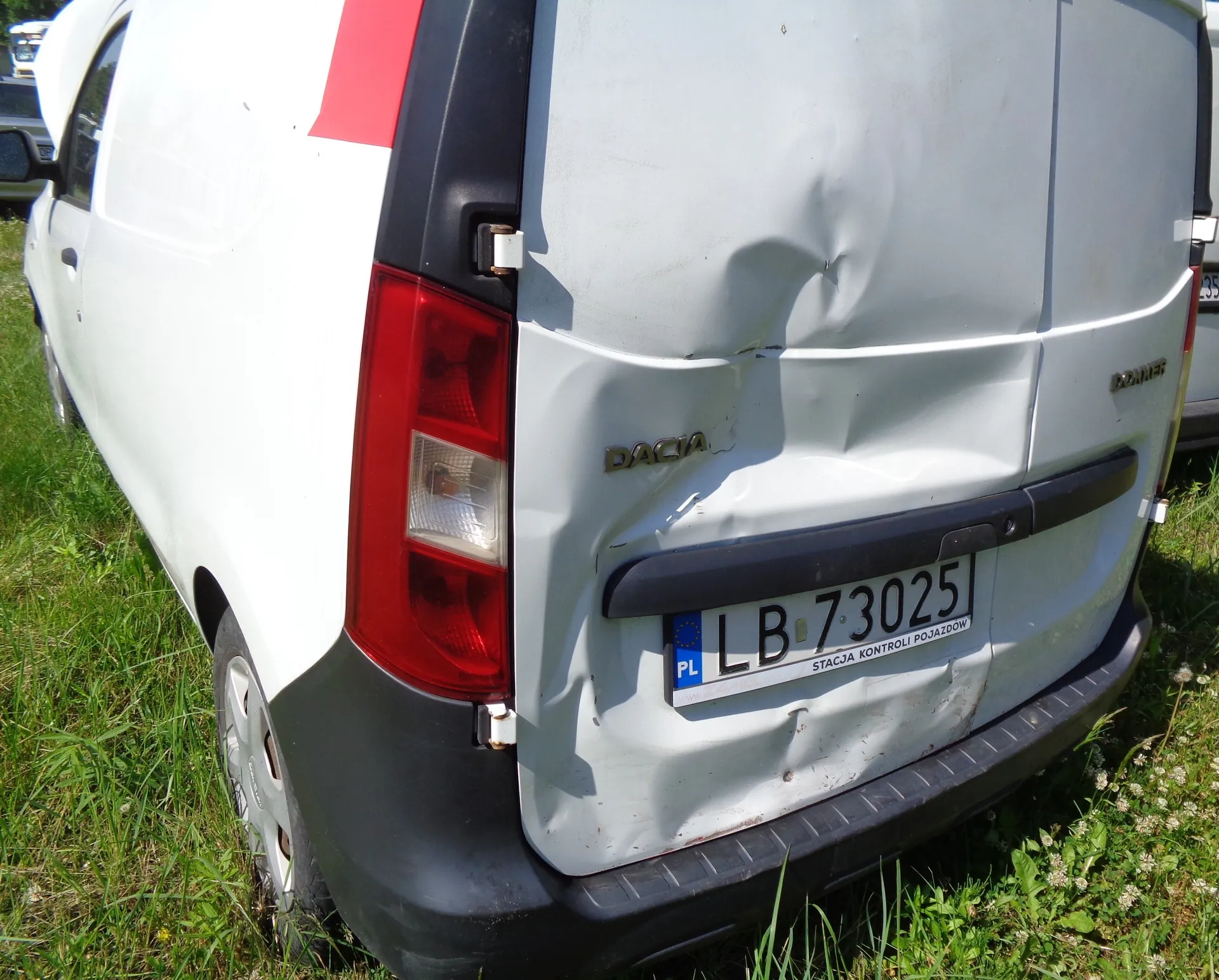 Tylna część białego samochodu marki Dacia z widocznym wgnieceniem na klapie bagażnika. Pojazd posiada polską tablicę rejestracyjną o numerze LB 73025, pod którą znajduje się napis 'STACJA KONTROLI POJAZDÓW'. Samochód stoi na trawie.