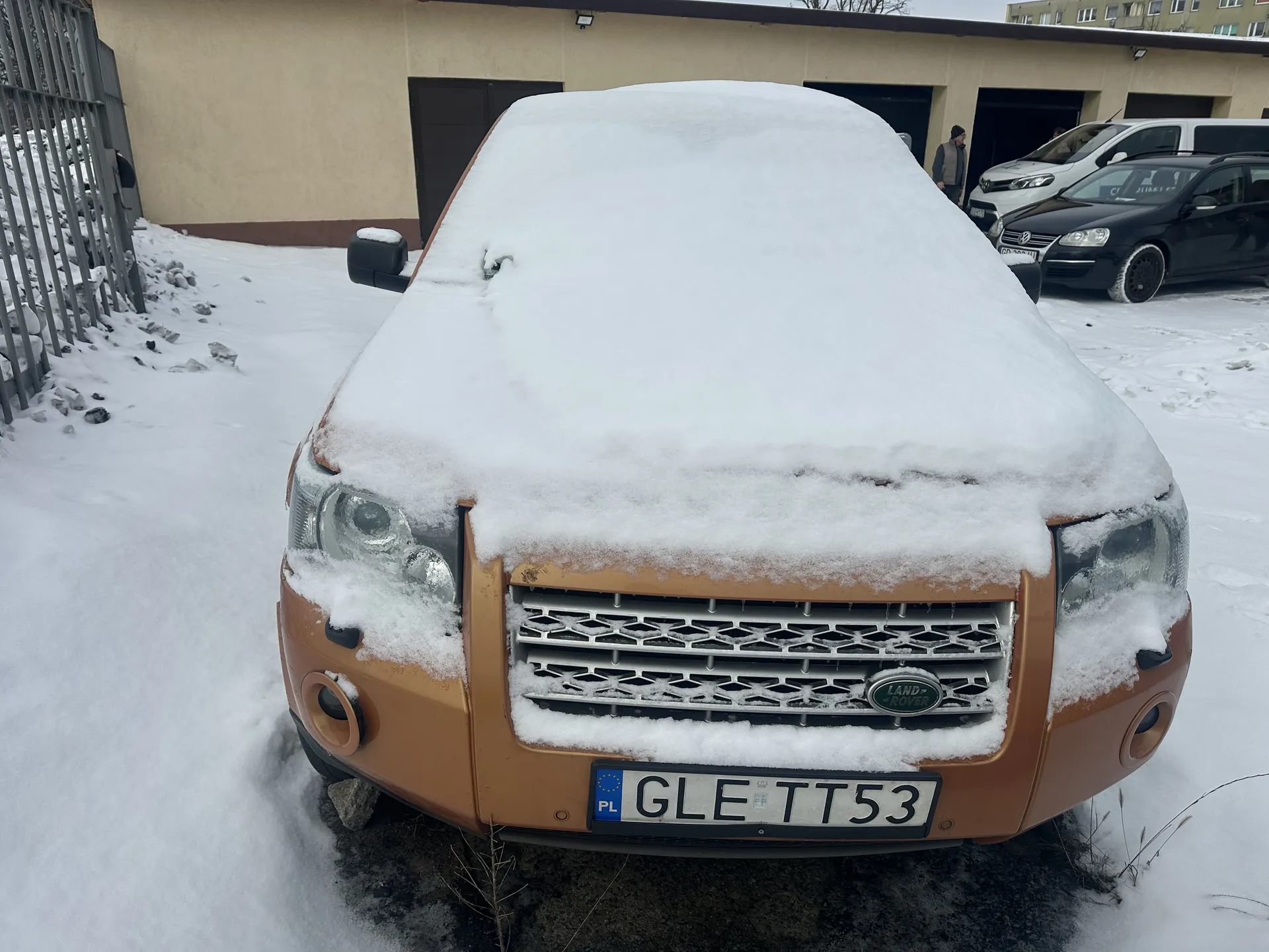 Przedni widok pomarańczowego samochodu marki Land Rover pokrytego grubą warstwą śniegu. Na masce widoczny jest logo marki, a na dole tablica rejestracyjna z polskim kodem PL i numerem GLE TT53. W tle budynek z bramami garażowymi, czarny i biały samochód oraz osoba stojąca blisko budynku. Cały obszar otoczony jest śniegiem, po lewej stronie metalowe ogrodzenie.
