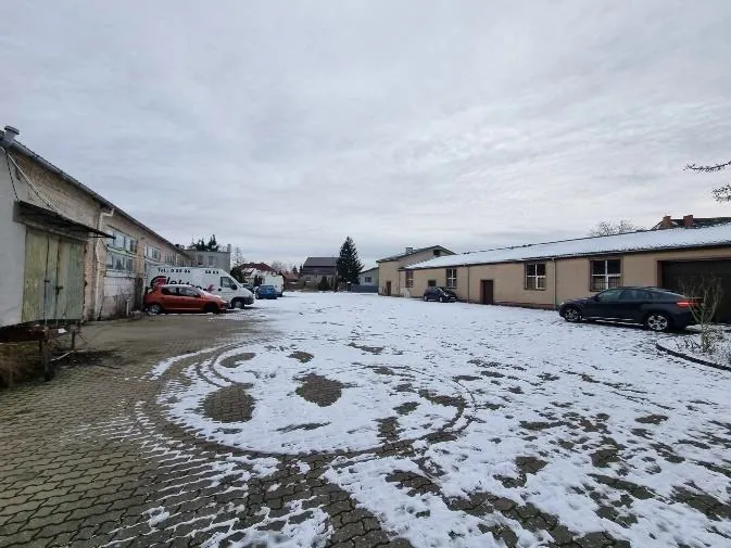 Zimowy dziedziniec z częściowo zasłanym śniegiem brukowanym podjazdem. Po lewej stronie stoją budynki z zielonymi bramami garażowymi, po prawej budynek o beżowej elewacji z oknami i garażem. Na dziedzińcu zaparkowane są samochody: czerwony, biały van, niebieski i czarny. Widoczne są ślady opon w śniegu. Tło stanowi pochmurne niebo i drzewa. Brak czytelnego tekstu.