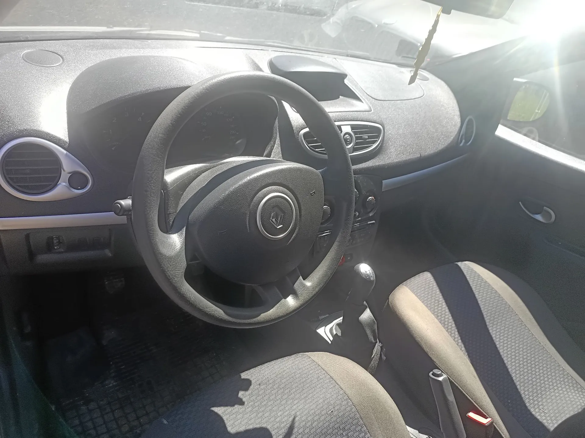 Wnętrze samochodu marki Renault widziane z perspektywy kierowcy. Widoczna jest czarna kierownica z logo Renault w centrum, deska rozdzielcza z licznikiem, nawiewami oraz dwa przednie fotele z ciemnoszarej tkaniny. Słońce wpada do wnętrza przez przednią szybę, tworząc jasne refleksy na powierzchniach.