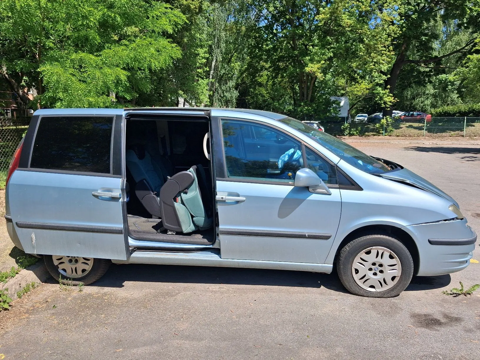 Srebrny samochód typu minivan zaparkowany na asfaltowym placu. Przesuwne drzwi po stronie pasażera są otwarte, ukazując wnętrze z jasnoniebieskimi fotelami. W tle widoczne są gęste, zielone drzewa oraz fragmenty innych zaparkowanych pojazdów za ogrodzeniem.