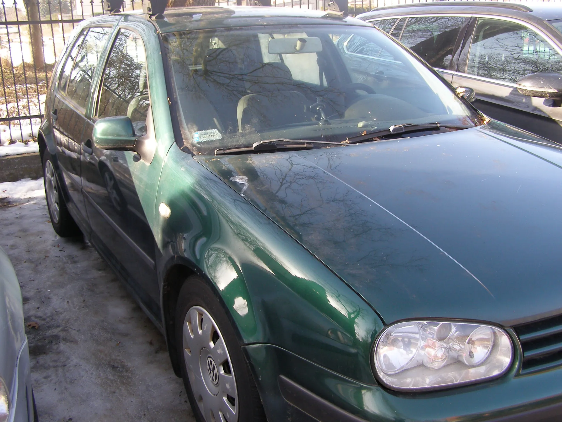 Zielony samochód osobowy marki Volkswagen, widok z przodu i boku, zaparkowany na obszarze z resztkami śniegu. W tle metalowe ogrodzenie i inne pojazdy. Na przedniej szybie widoczny naklejony znaczek. Samochód posiada srebrne felgi z logo marki.