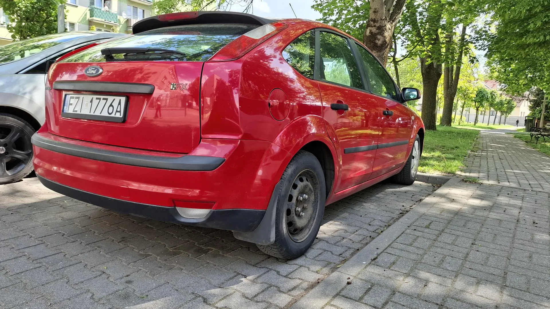 Czerwony samochód marki Ford Focus zaparkowany na kostce brukowej obok chodnika. Widoczny jest tył i prawy bok pojazdu. Samochód posiada polską tablicę rejestracyjną o numerze FZI 1776A. W tle widać zielone drzewa oraz fragment budynku mieszkalnego.