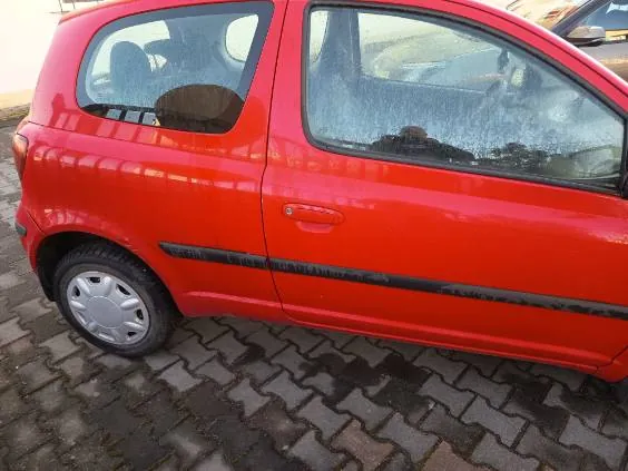 Czerwony samochód osobowy typu hatchback, widok z boku. Pojazd zaparkowany na brukowanej powierzchni z ciemnych i jasnych płytek. Widoczne są srebrne felgi, czarna listwa boczna oraz zabrudzone szyby. W tle fragment innego pojazdu i otoczenia.