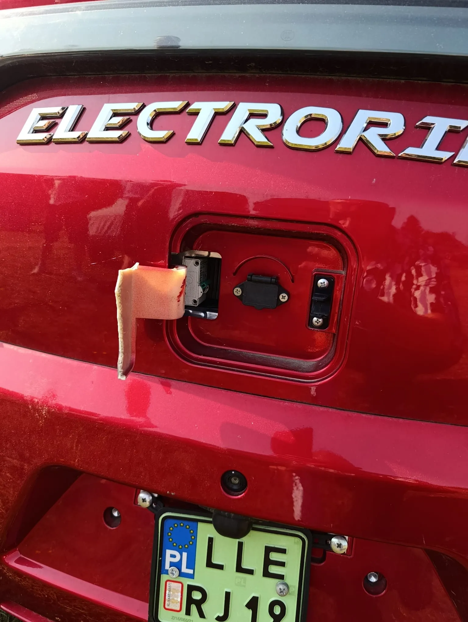 Zbliżenie na tylną część czerwonego samochodu. Na górze widnieje srebrny napis 'ELECTRORI'. Poniżej znajduje się otwarta klapka gniazda przyczepy, z której wystaje kawałek jasnego materiału lub taśmy. Na dole widoczna jest fragment tablicy rejestracyjnej z oznaczeniem kraju 'PL' oraz literami 'LLE' i 'RJ19'.