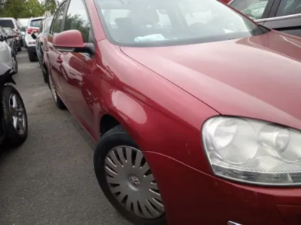 Czerwony samochód osobowy zaparkowany na placu parkingowym. W tle widoczne są inne pojazdy, w tym srebrny samochód po lewej stronie i białe auta w oddali. Samochód ma srebrne felgi i czerwone lusterko boczne. Asfaltowa powierzchnia parkingu oraz drzewa w tle.