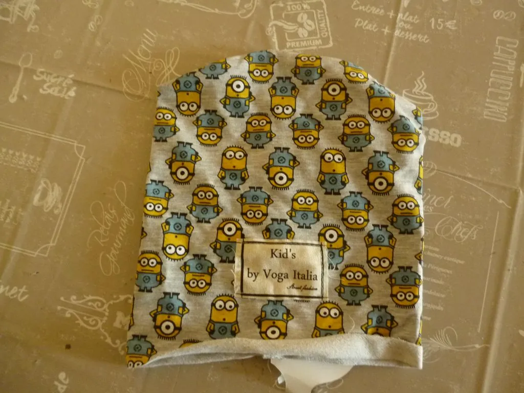 Szara czapka dziecięca z nadrukiem przedstawiającym żółte stworki przypominające Minionki w różnych pozach i z goglami. Na przodzie czapki znajduje się biała prostokątna metka z czarnym napisem: 'Kid's by Voga Italia'. Czapka leży na beżowym tle z białymi rysunkami i napisami w stylu kawiarnianym.