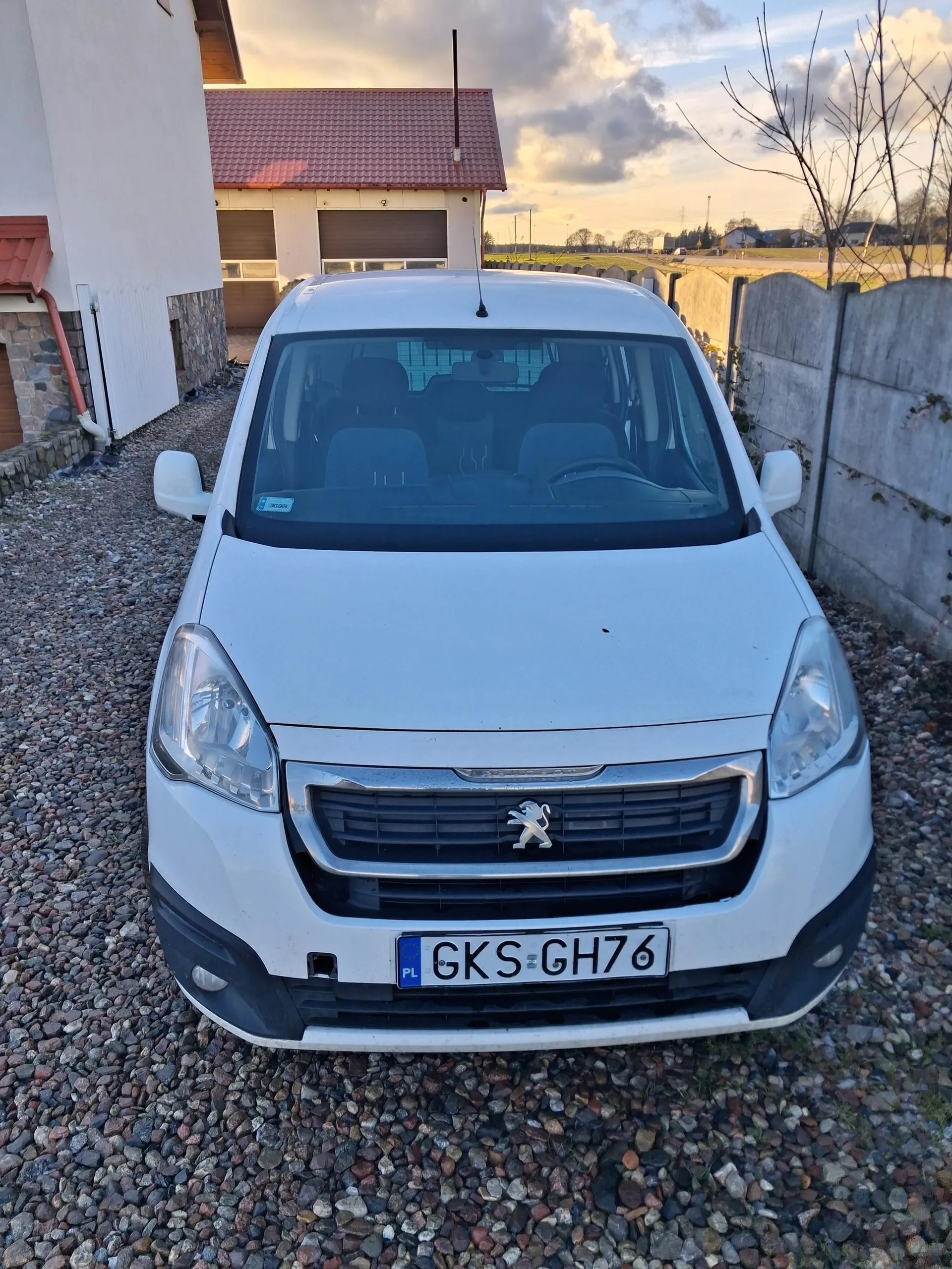 Biały samochód dostawczy marki Peugeot z tablicą rejestracyjną PL GKS GH76, zaparkowany na podwórku z kamieni. W tle budynek z czerwonym dachem, dwoma bramami garażowymi, betonowym ogrodzeniem po prawej stronie oraz drzewami bez liści. Niebo z chmurami, widoczne światło zachodzącego lub wschodzącego słońca.