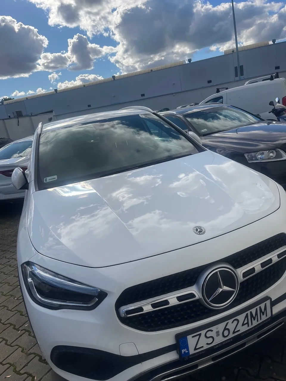 Biały samochód marki Mercedes-Benz zaparkowany na placu. Widoczny jest przód pojazdu z dużym logo gwiazdy na grillu oraz tablicą rejestracyjną o numerze ZS 624MM. W tle znajdują się inne samochody, biały budynek przemysłowy oraz niebo z białymi chmurami.