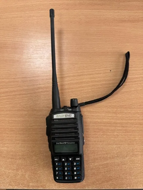 Czarna radiostacja marki Baofeng leżąca na drewnianym blacie. Urządzenie posiada długą antenę skierowaną w górę, klawiaturę numeryczną na dole oraz mały ekran LCD z napisem 'Dual Band FM Transceiver'. Z boku urządzenia odchodzi czarny sznurek do zawieszenia.