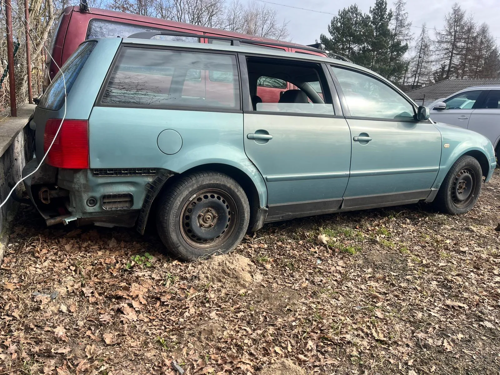 Niebieski samochód typu kombi z uszkodzonym tyłem, zaparkowany na podwórku pokrytym suchymi liśćmi. W tle czerwony van, srebrny samochód osobowy, drzewa bez liści oraz dom z dachem. Samochód ma brudne felgi, a tylna część wykazuje ślady uszkodzeń. Na ziemi widoczne są suche liście i niewielkie plamy trawy.