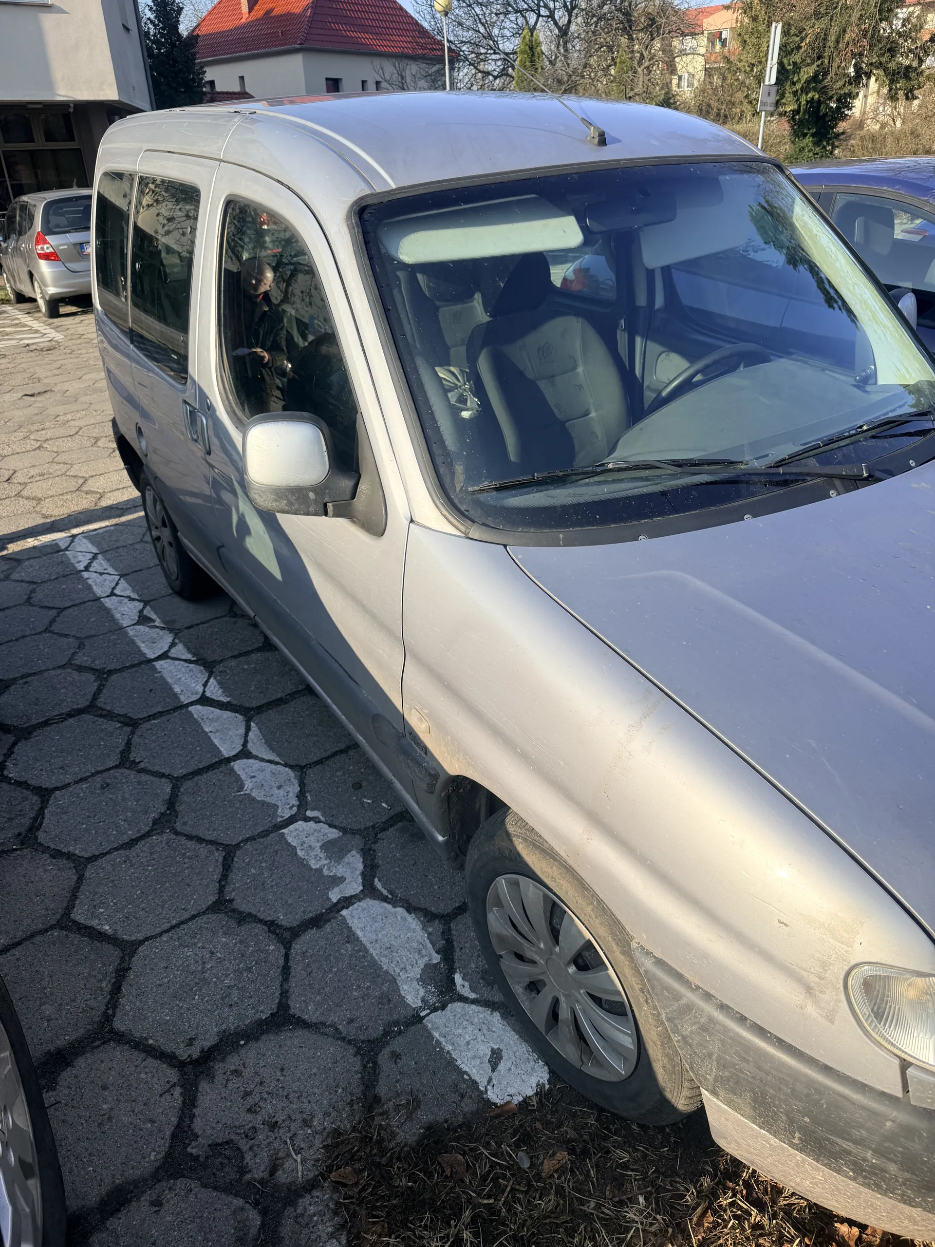 Srebrny samochód zaparkowany na brukowanej ulicy z sześciokątnymi kamieniami. Widoczne przednie szyby, wnętrze z siedzeniami, koła z felgami. W tle budynki z czerwonymi dachami, drzewa bez liści i znak drogowy. Po prawej stronie sucha trawa i liście. Samochód znajduje się pod kątem, a horyzont jest pionowy.