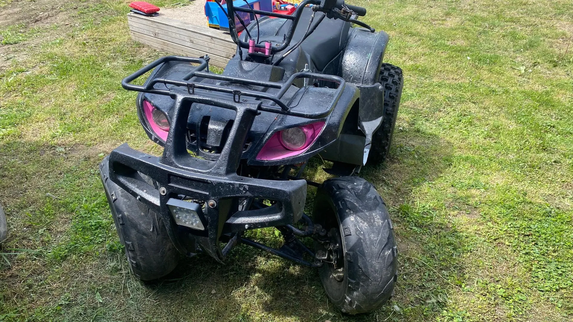 Czarny quad zaparkowany na trawiastym terenie. Pojazd posiada metalowy bagażnik z przodu, różowe detale wokół reflektorów oraz szerokie, terenowe opony. W tle widoczny jest drewniany podest z kolorowymi zabawkami.