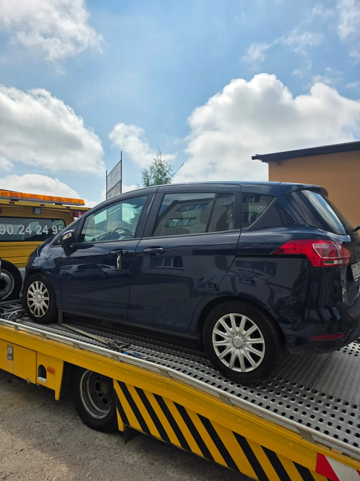 Ciemnoniebieski samochód osobowy typu hatchback zaparkowany na żółtej platformie lawety. Laweta posiada czarno-żółte pasy ostrzegawcze na bocznej krawędzi. W tle widoczne jest jasne niebo z białymi chmurami oraz fragment żółtego pojazdu z numerem telefonu 90 24 24 9. Po prawej stronie znajduje się fragment beżowego budynku.