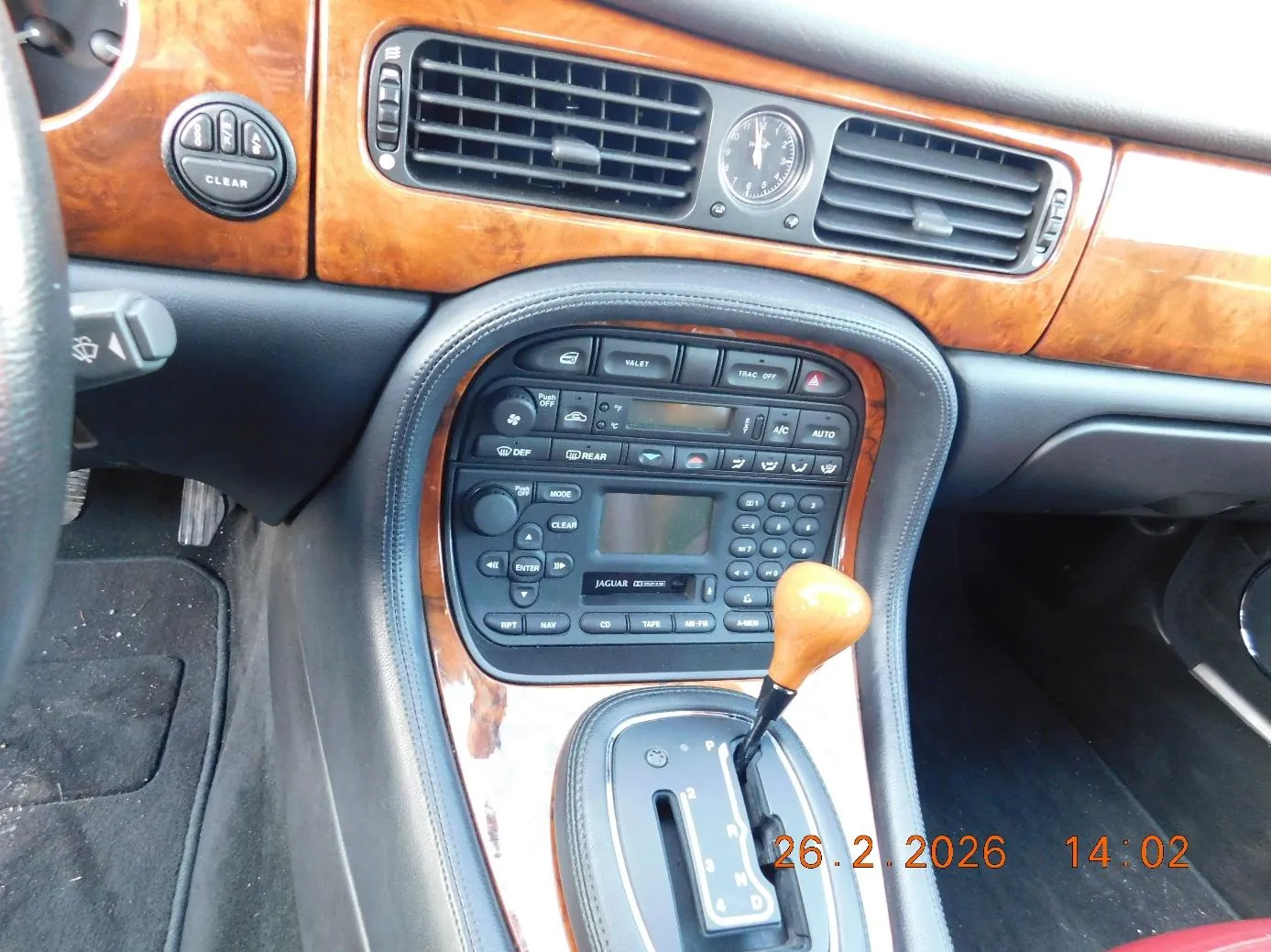 Wnętrze samochodu z widoczną deską rozdzielczą i centralną konsolą. Deska wykończona jest drewnianą lamelą i czarną tapicerką. W górnej części znajdują się czarne klimatyzacyjne wywiewacze, analogowy zegar oraz przyciski z napisem CLEAR. W centralnej części panelu widoczne są przyciski sterujące (VALET, TRAC OFF, A/C, AUTO), ekran, klawiatura oraz napis JAGUAR na urządzeniu audio. Poniżej znajduje się dźwignia zmiany biegów z drewnianą rączką oraz oznaczeniami P, R, N, D, 1-4. W prawym dolnym rogu obrazu widoczny jest pomarańczowy znaczek daty i godziny: 26.2.2026 14:02.