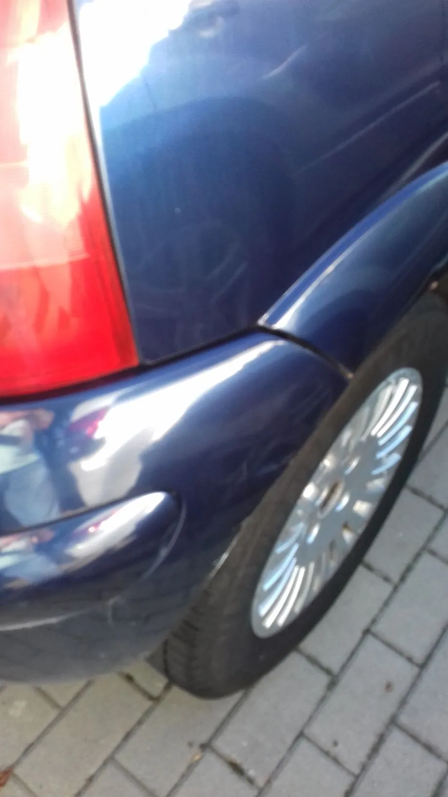 Zbliżenie na tylną część niebieskiego samochodu, obejmujące fragment czerwonego tylnego światła, błotnik oraz koło z wieloramienną srebrną felgą, zaparkowane na kostce brukowej.