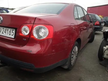 Czerwony samochód osobowy marki Volkswagen z tablicą rejestracyjną J24165, widok od tyłu i boku. Samochód stoi na asfaltowym parkingu, w tle widoczne są inne pojazdy oraz czerwony budynek. Na tylnym zderzaku i błotniku widoczne są drobne uszkodzenia.