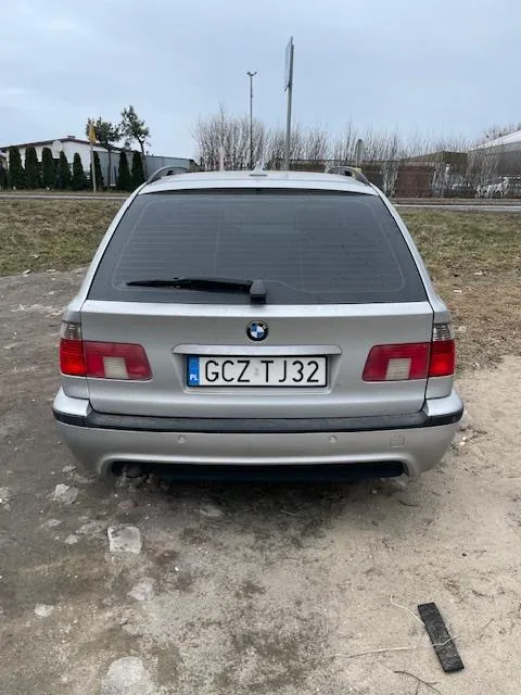 Tył srebrnego samochodu marki BMW z tablicą rejestracyjną GCZ TJ32. Samochód stoi na nierównym, częściowo zagruntowanym terenie. W tle widoczne są suche drzewa, budynki i słup. Na ziemi leży czarny przedmiot przypominający pilot. Niebo jest zachmurzone.