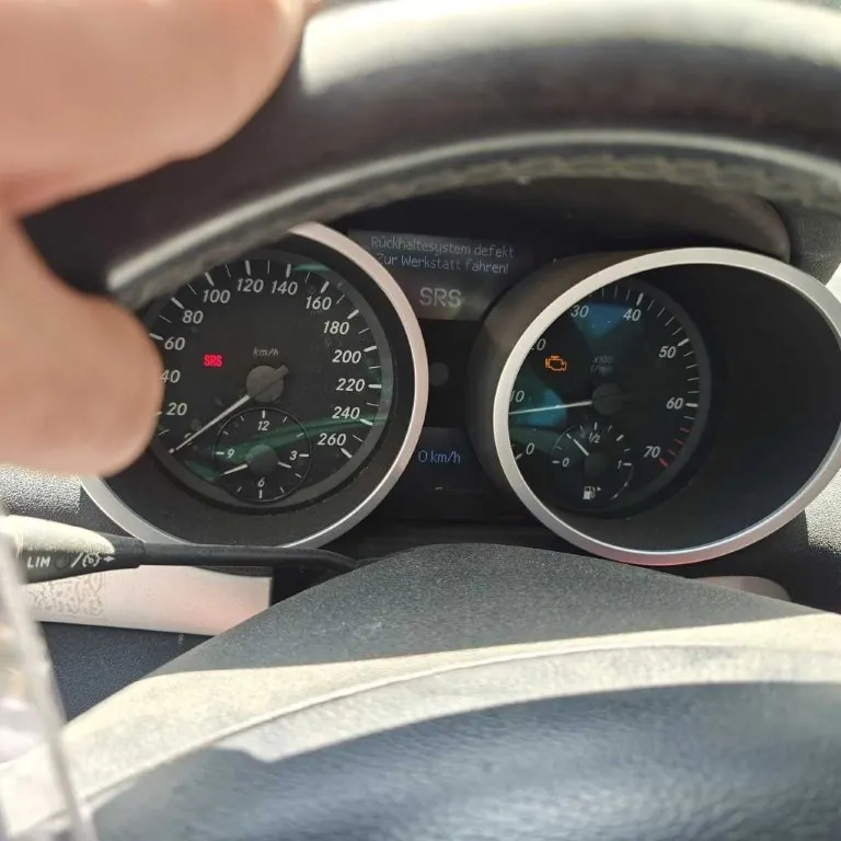 Zbliżenie na deski rozdzielcze samochodu z widocznym licznikiem prędkości (0-260 km/h) i obrotomierzem. Na liczniku prędkości widoczny czerwony napis SRS oraz wskaźnik prędkości 0 km/h. W centralnej części wyświetlacza komunikat w języku niemieckim: Rückhaltesystem defekt. Zur Werkstatt fahren! oraz napis SRS. Po prawej stronie obrotomierz z pomarańczowym symbolem ostrzegawczym. W lewym górnym rogu widoczna część dłoni trzymającej kierownicę. W dolnej części obrazu fragment kierownicy i elementów wnętrza pojazdu.
