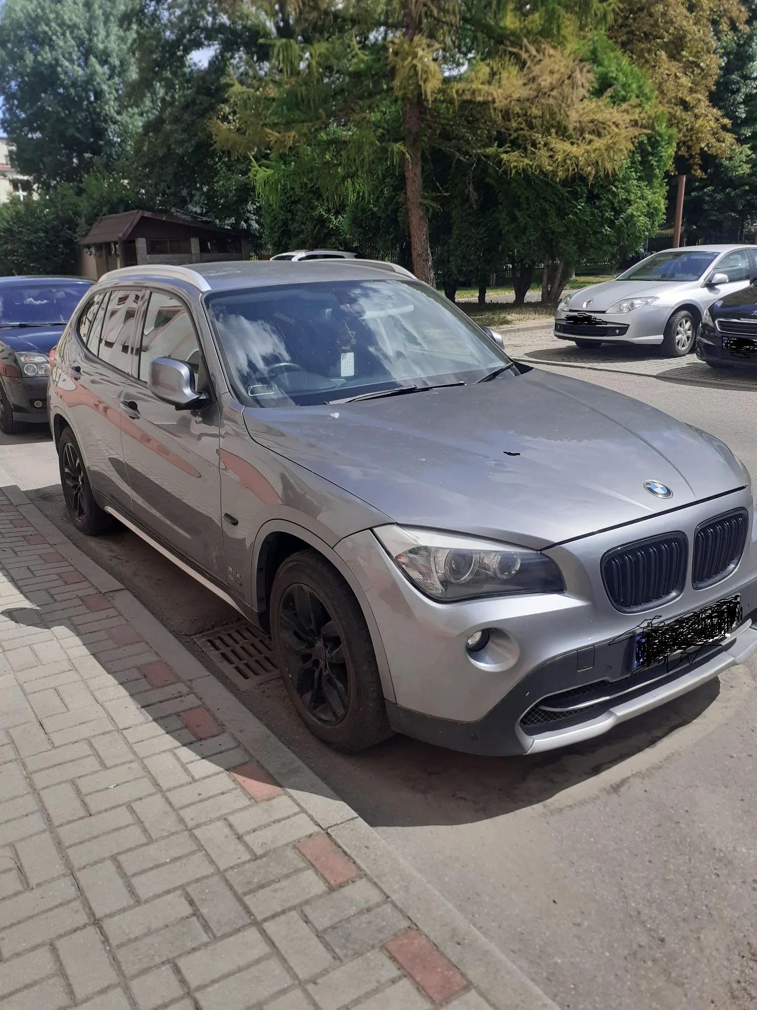 Srebrny samochód marki BMW X3 zaparkowany na ulicy obok chodnika z kostki brukowej. W tle widoczne są zielone drzewa, fragment budynku oraz inny srebrny samochód zaparkowany dalej.