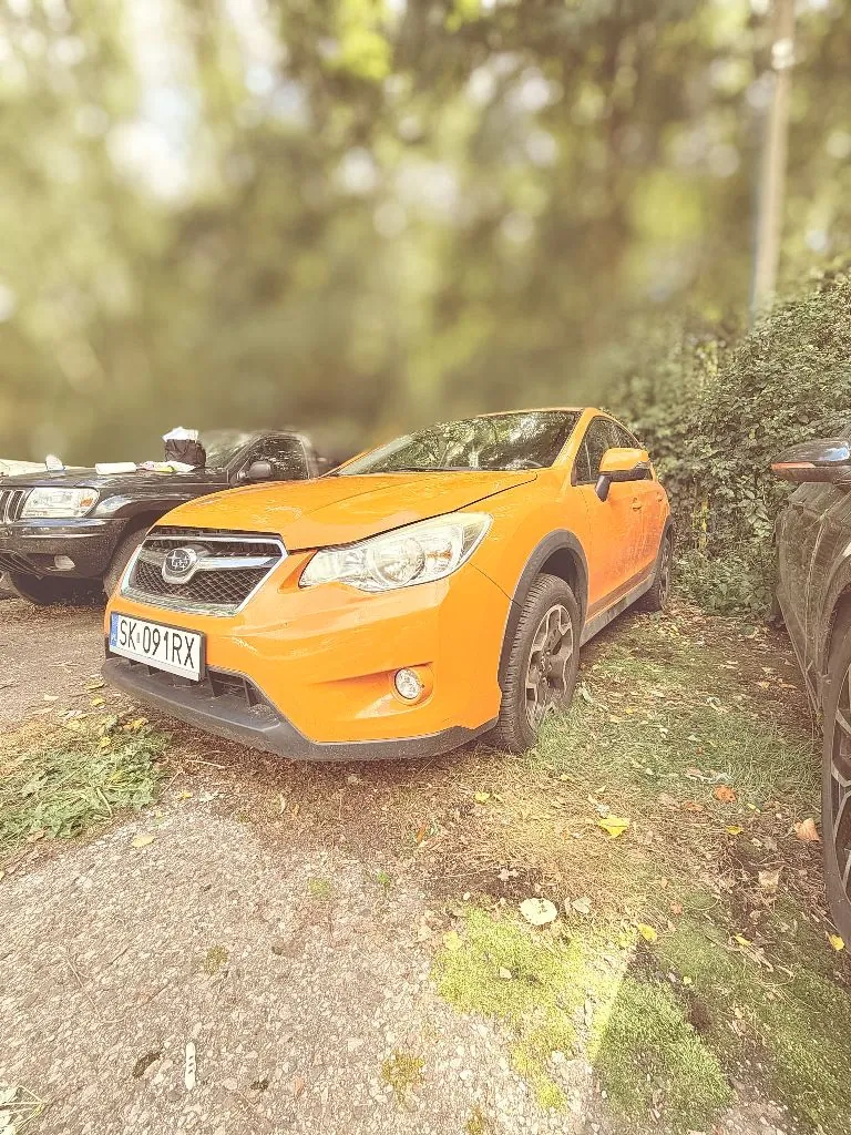 Pomarańczowy samochód marki Subaru XV zaparkowany na poboczu drogi, obok zielonych krzewów. Samochód ma polską tablicę rejestracyjną o numerze SK 091RX. W tle widoczny jest fragment innego, ciemnego samochodu oraz rozmyte drzewa.