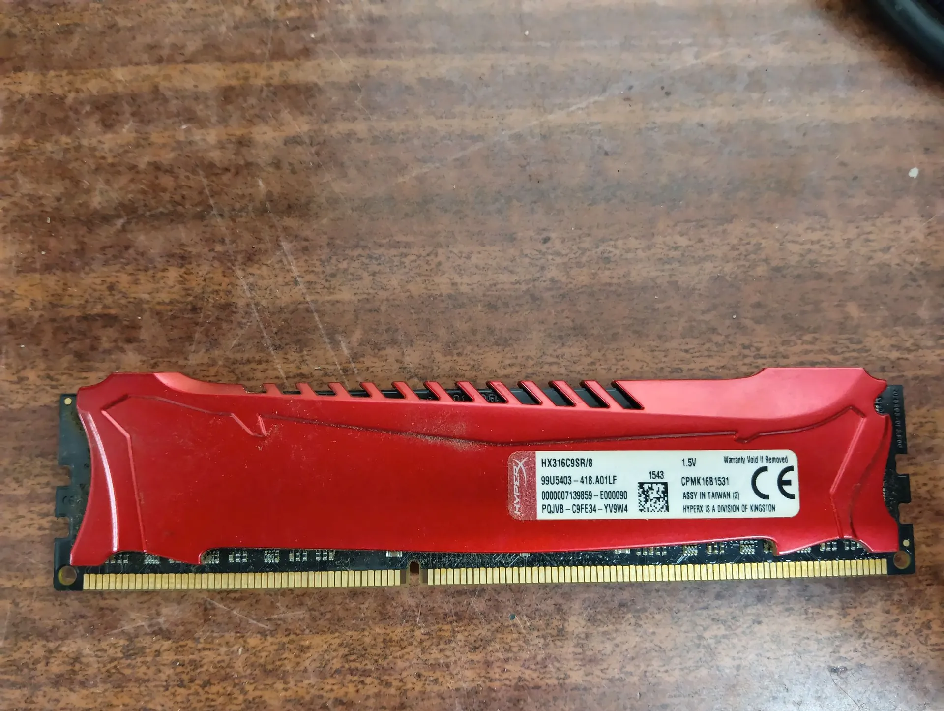 Moduł pamięci RAM z czerwonym radiatorem, leżący poziomo na drewnianym blacie. Na naklejce widnieją napisy: 'HX316C9SR/8', '1.5V', '99U5403-418.A01LF', '1543', 'CPMK1681531', '00000071389SR-E000090', 'ASSY IN TAIWAN (2)', 'PQJV8-C9FE34-YV9W4', 'HYPERX IS A DIVISION OF KINGSTON' oraz znak CE.