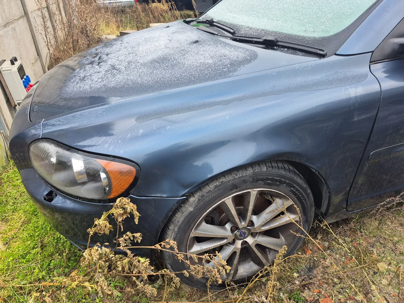 Ciemnoniebieski samochód z widoczną warstwą szronu na masce i przedniej szybie. Widoczne przednie lewe koło z aluminiową felgą, reflektor z pomarańczowym kierunkowskazem. Wokół samochodu sucha trawa i zarośla, po lewej stronie betonowa konstrukcja. Podłoże z trawą i ziemią.