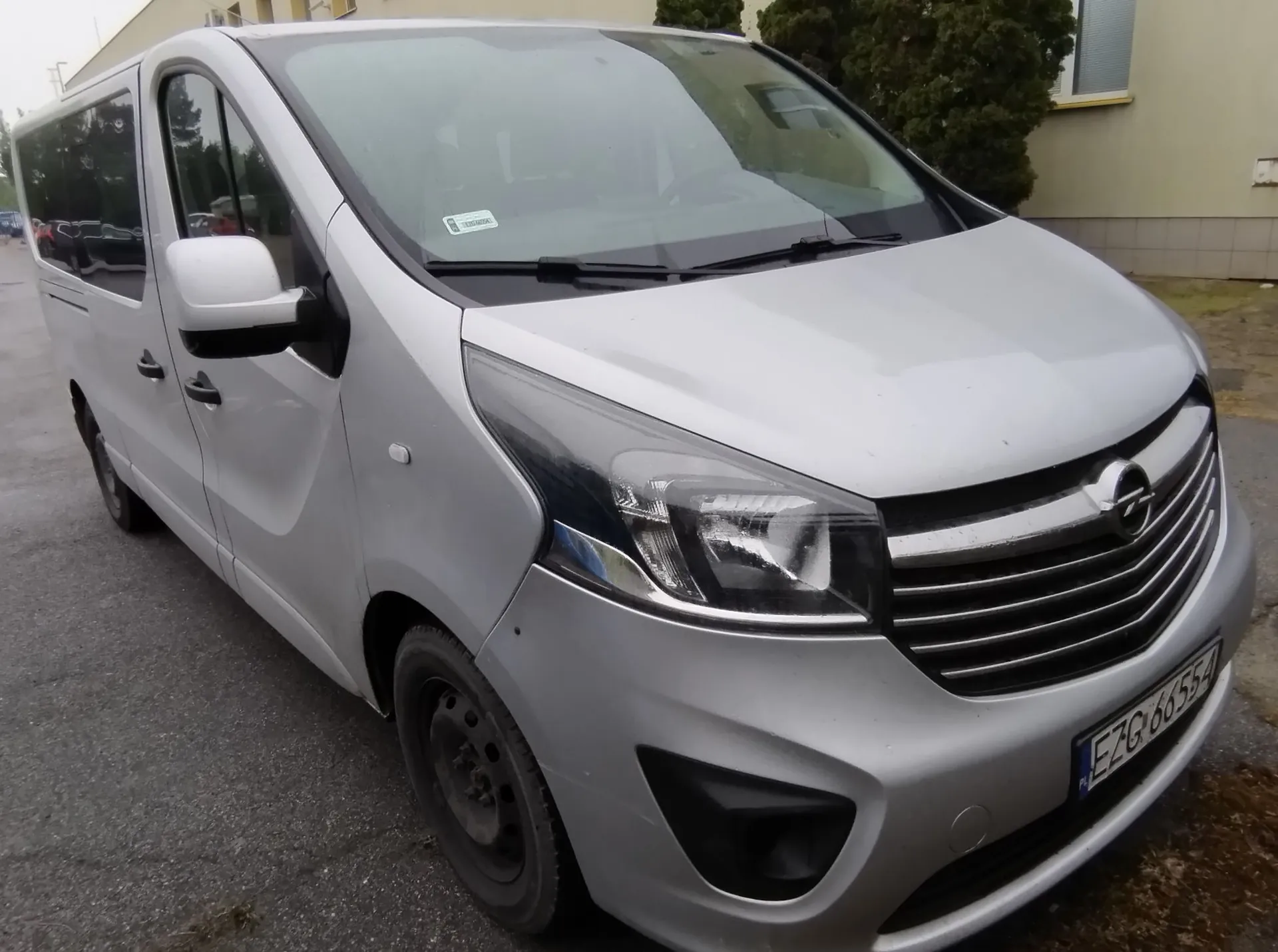 Srebrny samochód typu van marki Opel zaparkowany na asfaltowej nawierzchni. Widoczny jest przód i prawy bok pojazdu. Samochód posiada tablicę rejestracyjną o numerze EZG 66554. W tle widać fragment budynku oraz zielony krzew.