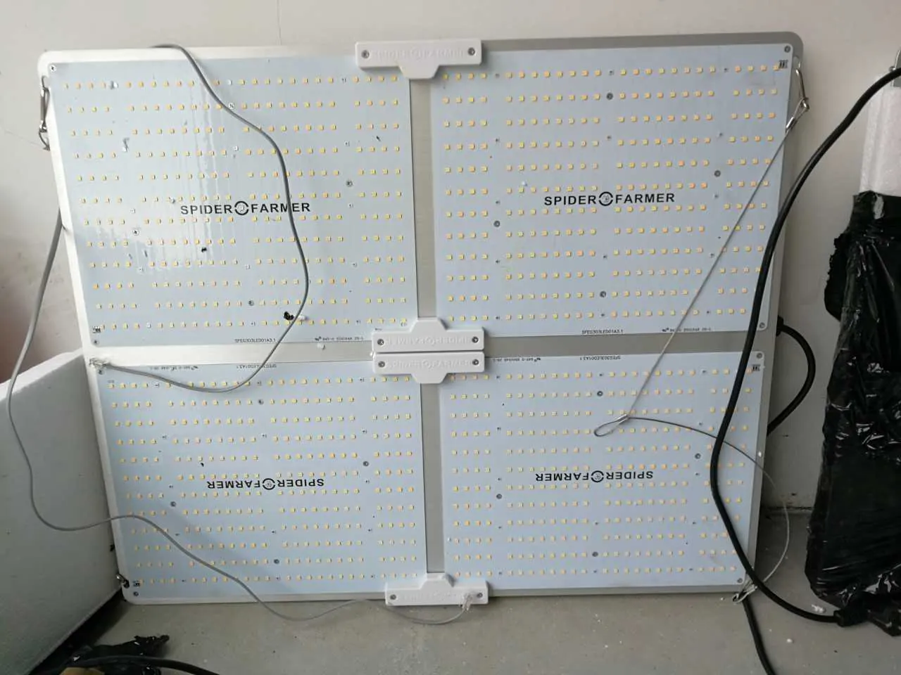 Cztery kwadratowe panele LED ułożone w siatce 2x2. Dwa górne panele mają napis 'SPIDER FARMER' w prawidłowej orientacji, dwa dolne w orientacji odwróconej. Panele są jasnoniebieskie z licznymi małymi żółtymi diodami LED. Widoczne są przewody elektryczne (czarne i szare) podłączone do paneli. Tło stanowi jasna ściana i szara podłoga. Po prawej stronie znajduje się czarna torba z plastiku.