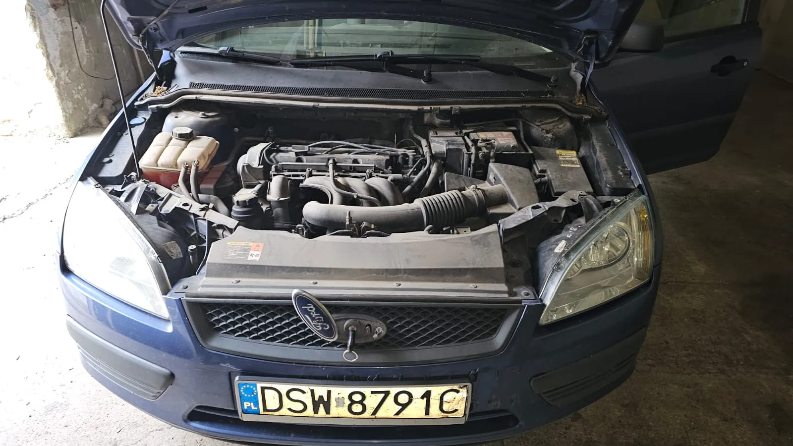 Widok z przodu na niebieski samochód marki Ford z otwartą maską, odsłaniającą silnik i elementy komory silnika. Na przednim zderzaku znajduje się tablica rejestracyjna o numerze DSW 8791C.