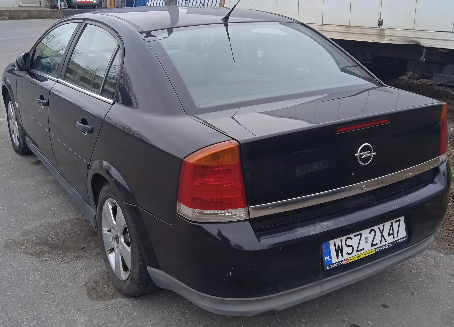 Czarny samochód osobowy marki Opel, widok z tyłu. Widoczna tablica rejestracyjna z polskim kodem PL i numerem WSZ 2X47. Na tablicy dodatkowe napisy: AUTO KOMIS, numer telefonu oraz informacja o ubezpieczeniu. Samochód zaparkowany na asfaltowej powierzchni, w tle fragment dużego pojazdu i budynku. Czerwone światła tylnego oświetlenia, srebrne felgi, czarna obudowa z logo Opel na bagażniku.